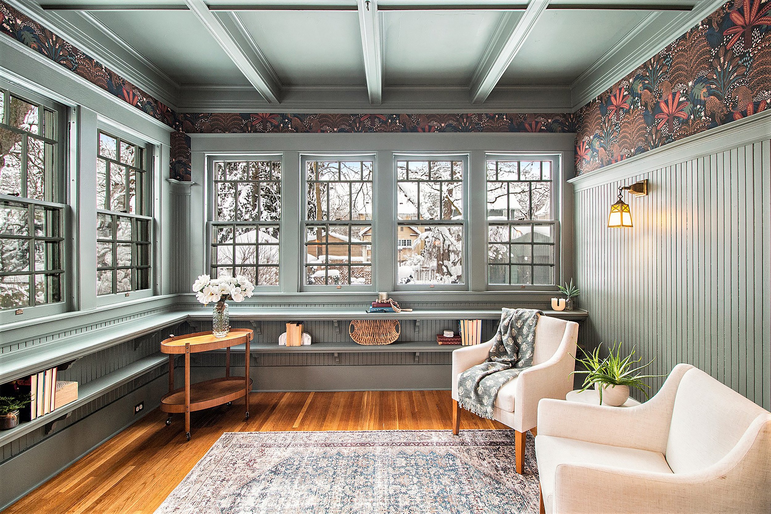 Living room with large windows showing snow outside, light green paneled walls, beige armchairs, a small wooden table with flowers, a bookshelf below the windows, a patterned rug, and a wall sconce light.