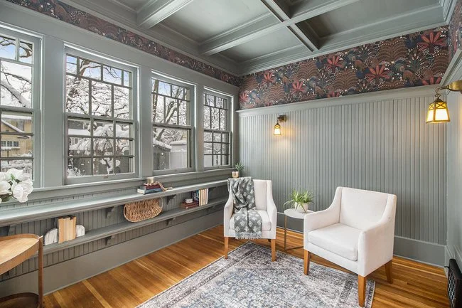 Cozy sunlit living room with large windows, white armchairs, a side table with a plant, built-in bookshelf, floral wallpaper, and hardwood flooring.