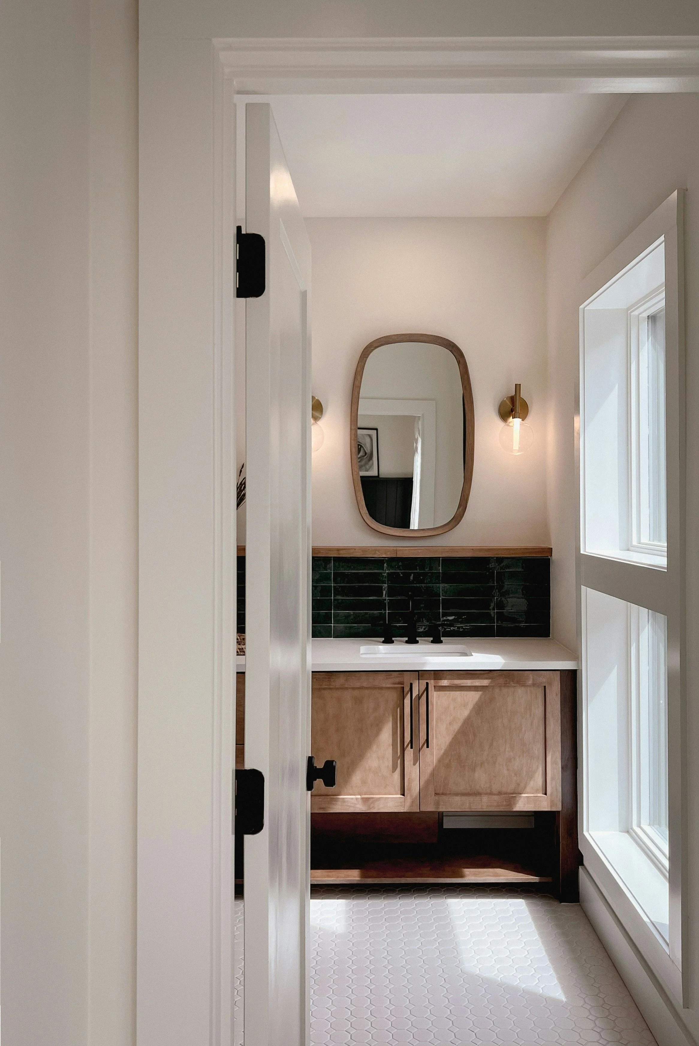 A small bathroom with a white door partially open, revealing a vanity with a wooden cabinet, a white sink, a mirror with a wooden frame, and two wall-mounted light fixtures. There is a window on the right side with sunlight streaming in.