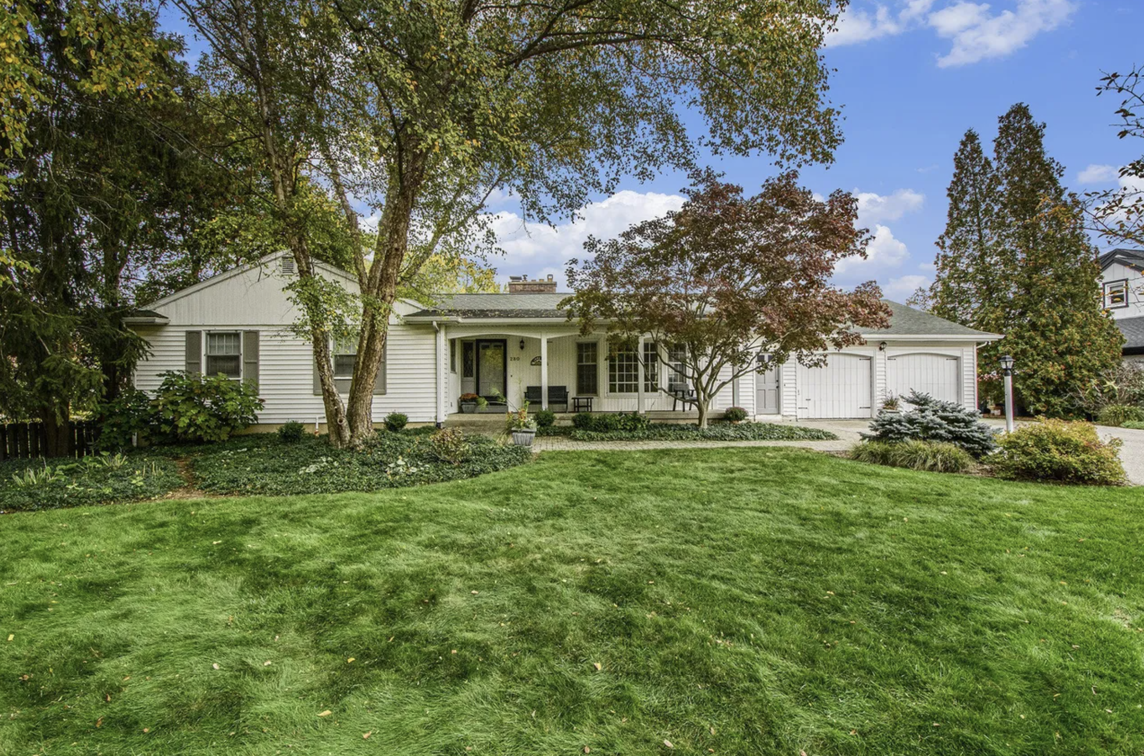 Front view of a white house with a manicured lawn, trees, and shrubs under a partly cloudy sky.