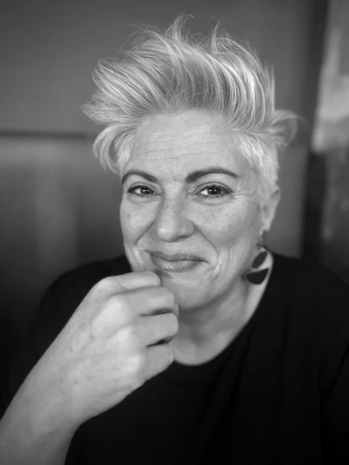 A woman with short, styled gray hair, smiling softly with her hand near her chin, wearing a dark top and an earring, in a black and white photo.