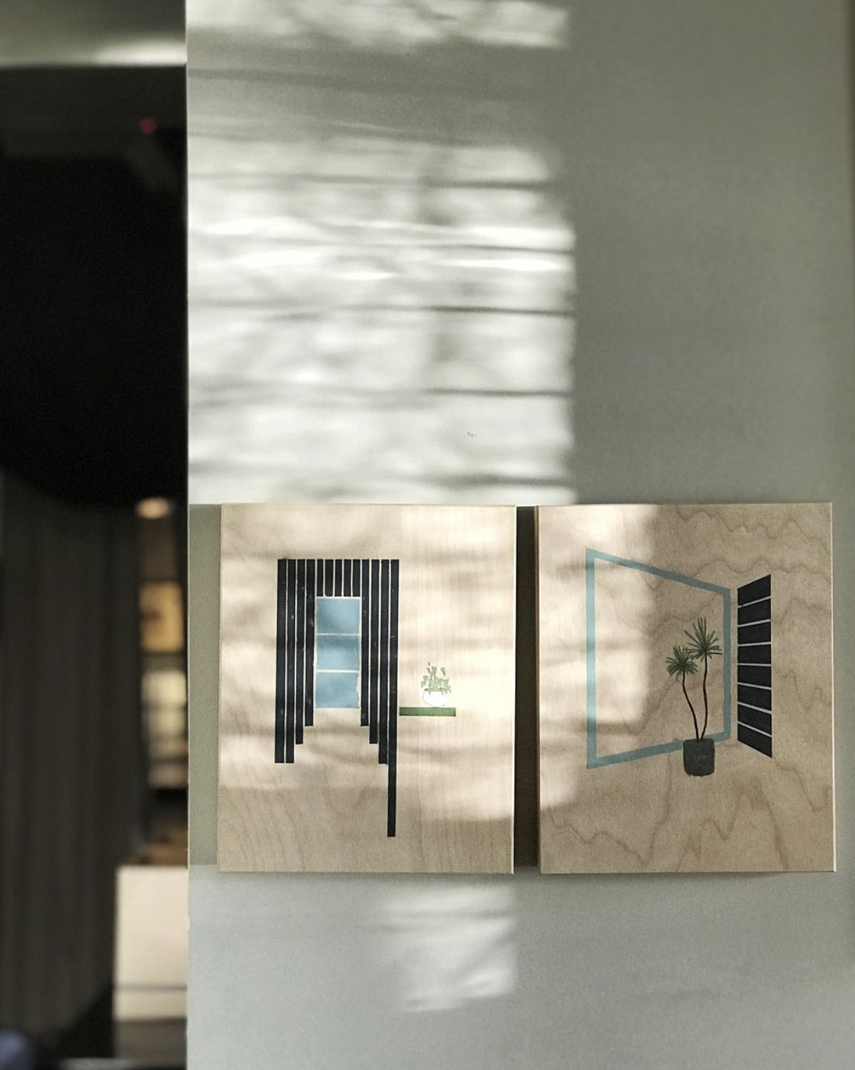 Two minimalist drawings of open windows with plants on the windowsills, displayed on wooden panels, with sunlight and shadow patterns on the wall behind.