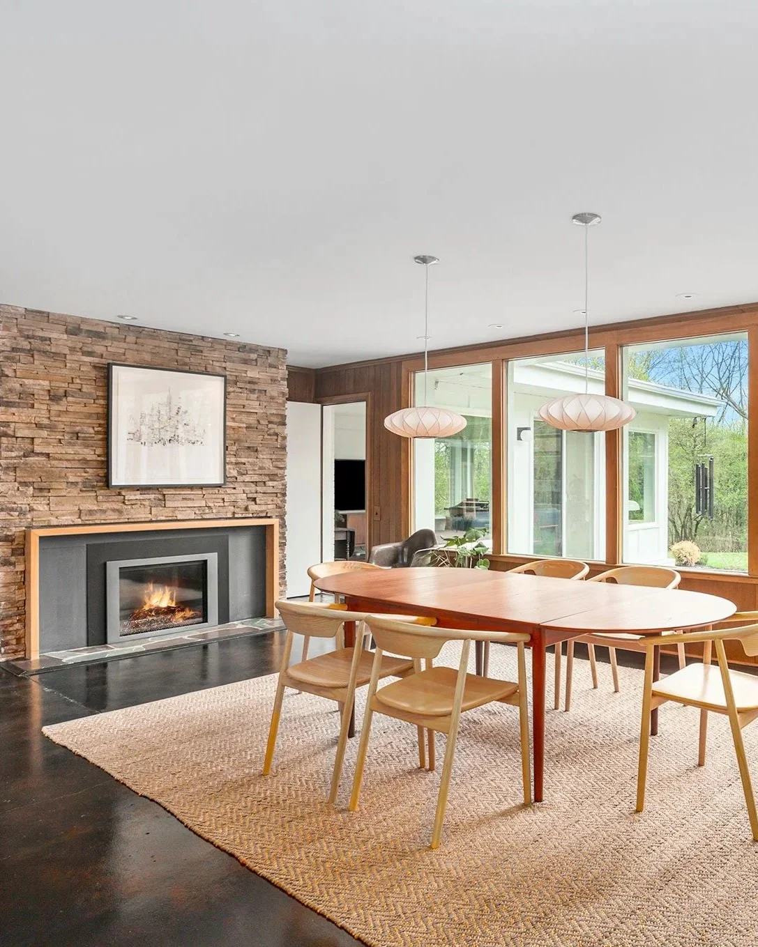 Dining room with large wooden table, light wood chairs, a stone fireplace, large windows, and pendant lights.