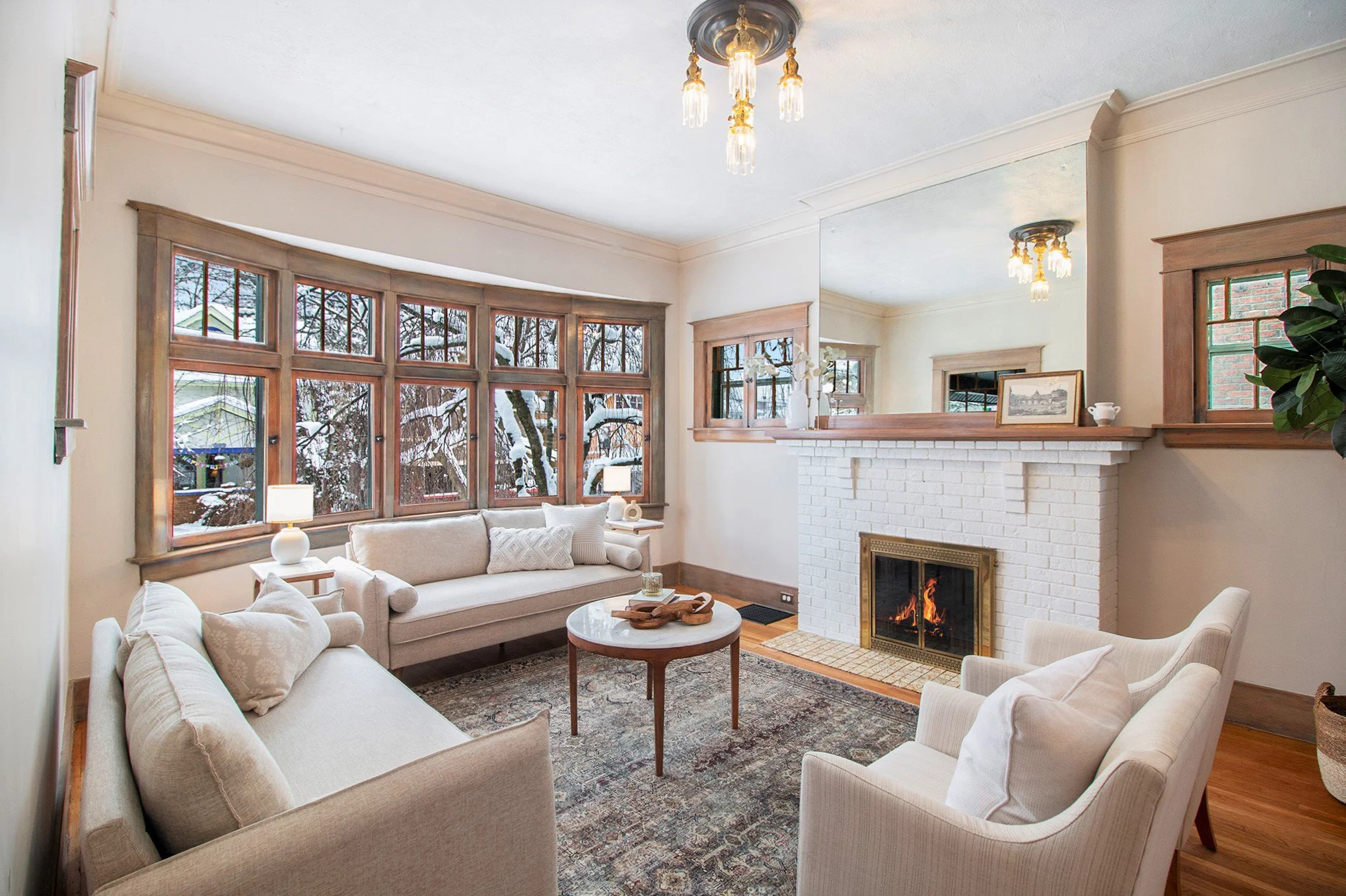 Living room with large bay window, white brick fireplace, beige sofas, and wooden accents, with snow outside.