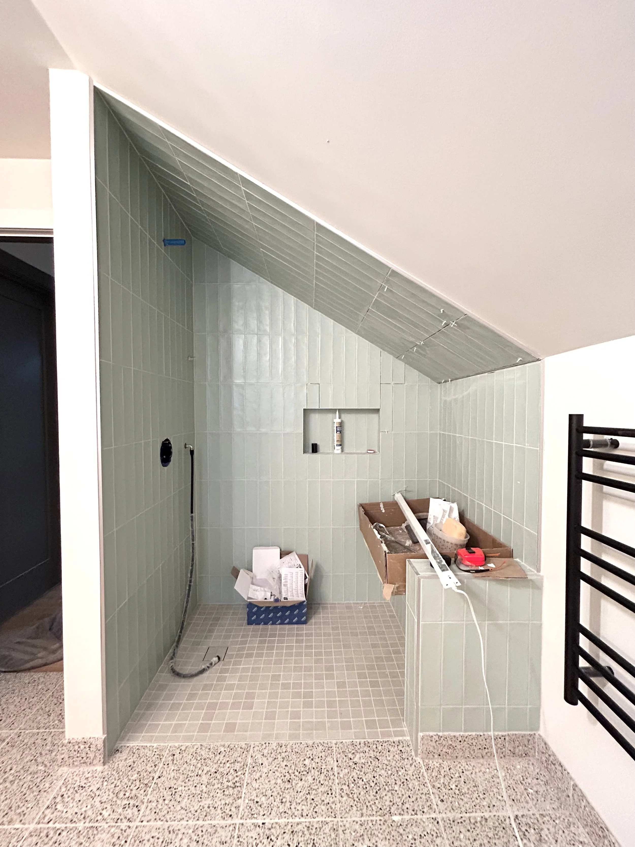 A bathroom under construction with green tiled walls, a small built-in shelf, and construction tools and materials on the floor.