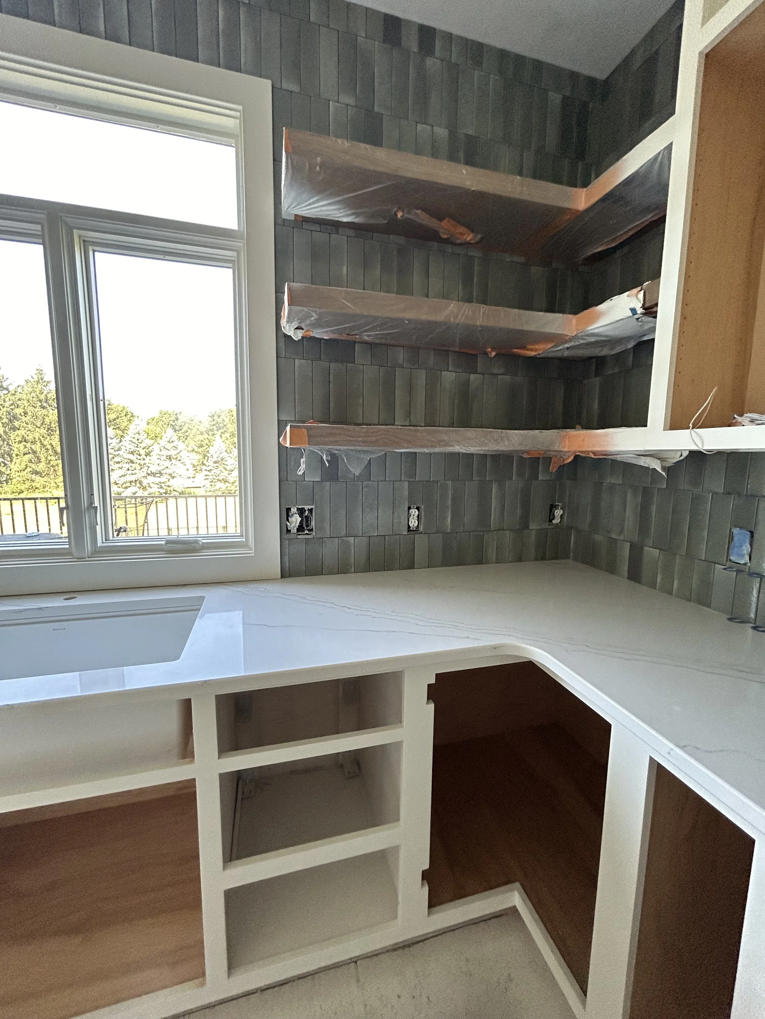 A kitchen under renovation with white cabinets, a marble countertop, a window showing an outdoor view, and dark tiled backsplash. There are shelves wrapped in protective plastic on the wall, and electrical outlets and plumbing for appliances.