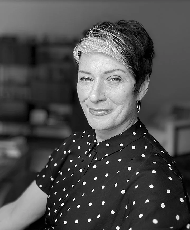 A woman with short hair styled with highlights, wearing a black top with white polka dots, smiling at the camera in an indoor setting.