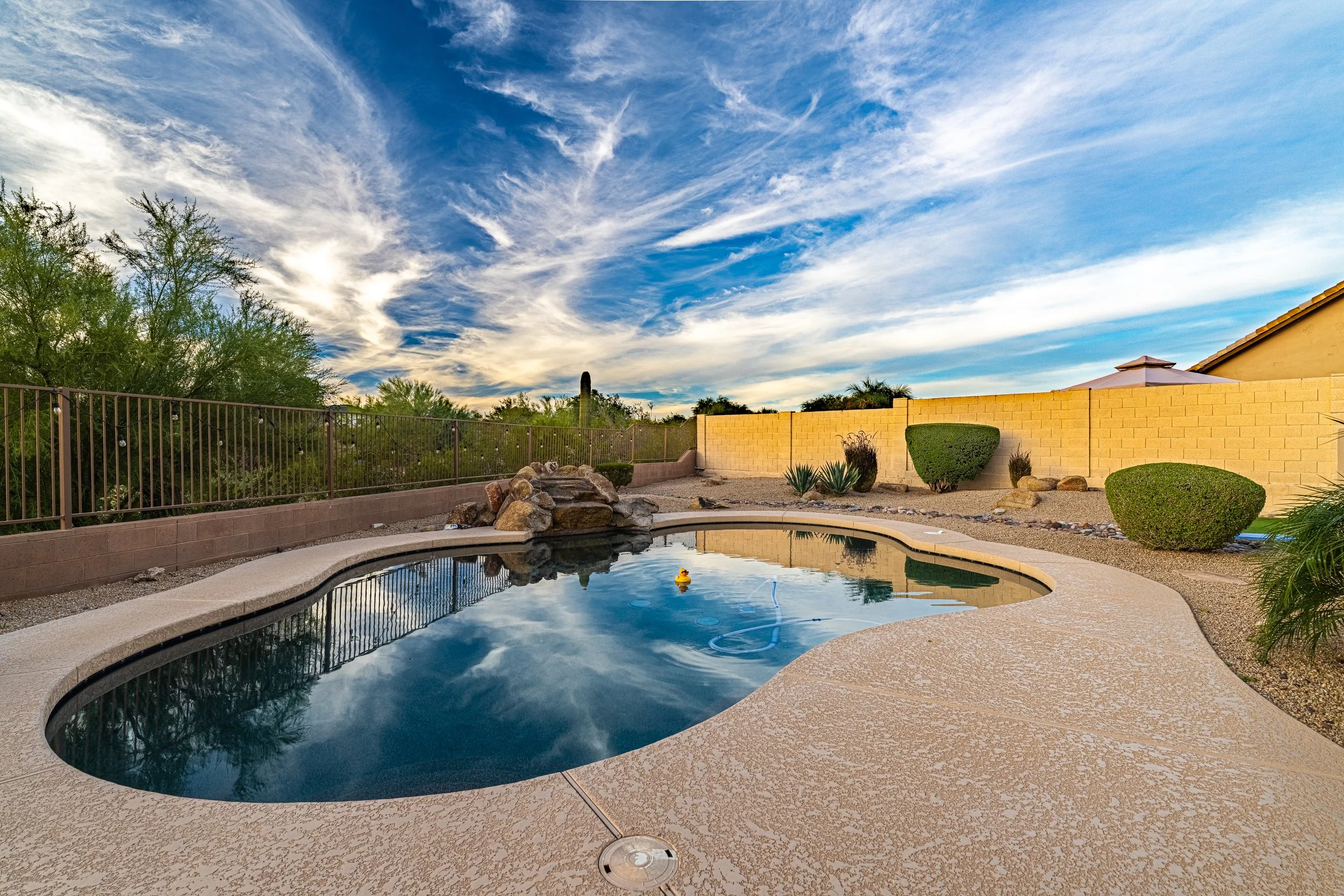 A backyard swimming pool with a natural rock waterfall feature, surrounded by desert plants, a brick fence, and a cloudy blue sky.