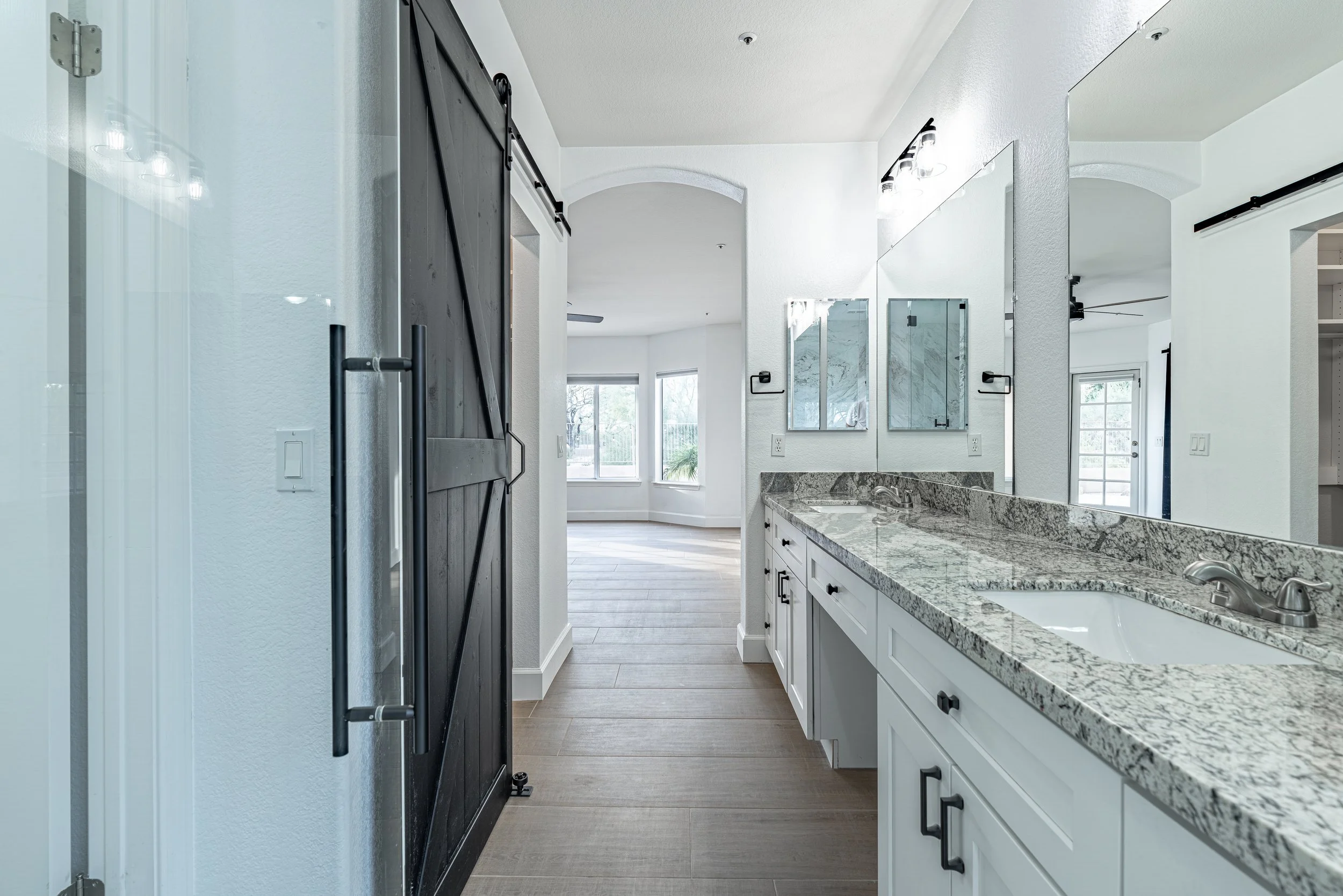 Bathroom with white cabinets, granite countertop, large mirror, and two sinks. Barn door on the left, living area with large windows in the background.