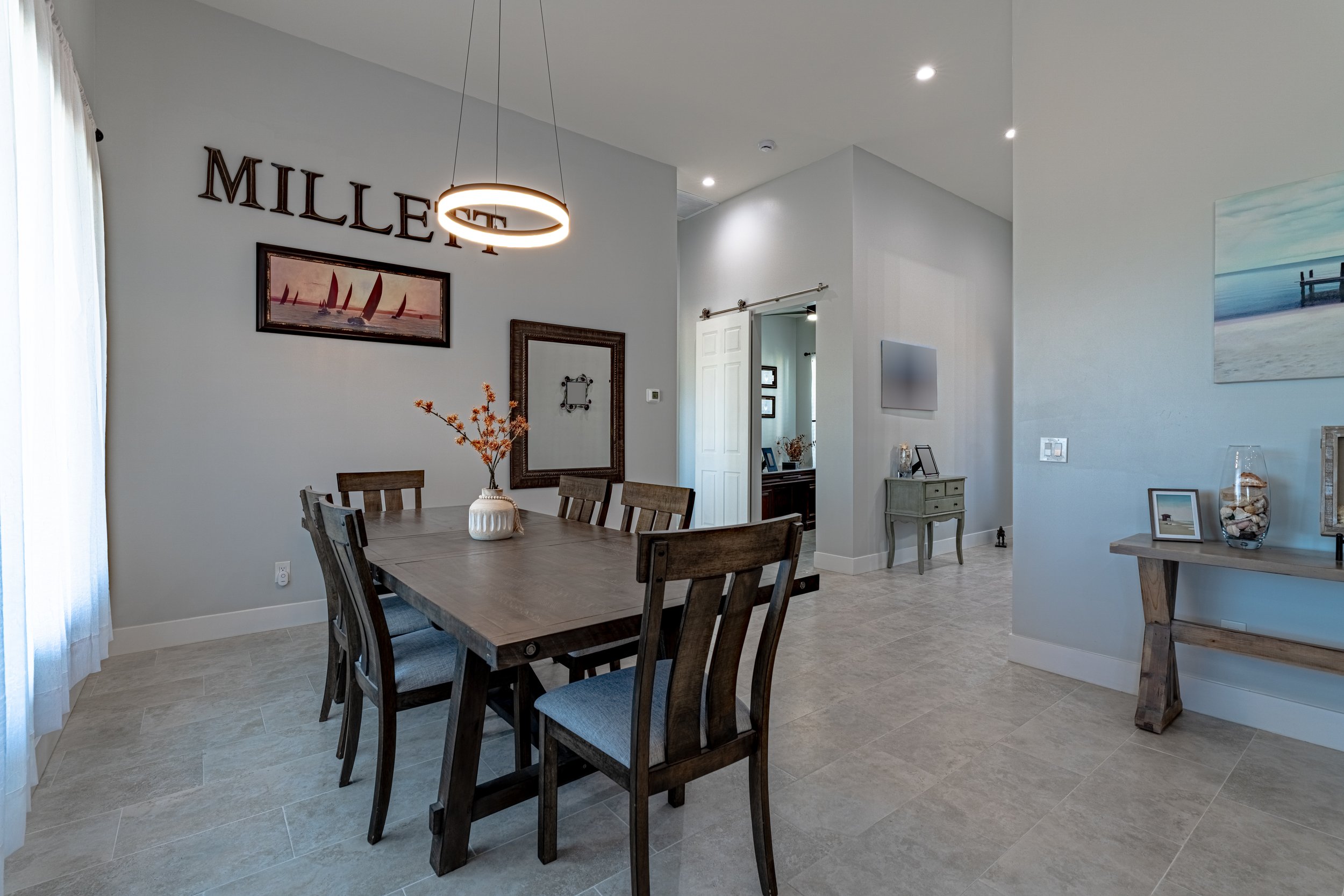Dining room with wooden table and chairs, wall decorations including an art piece of sailboats, a framed mirror, a round hanging light fixture, and a side table with a picture and a glass jar with shells.