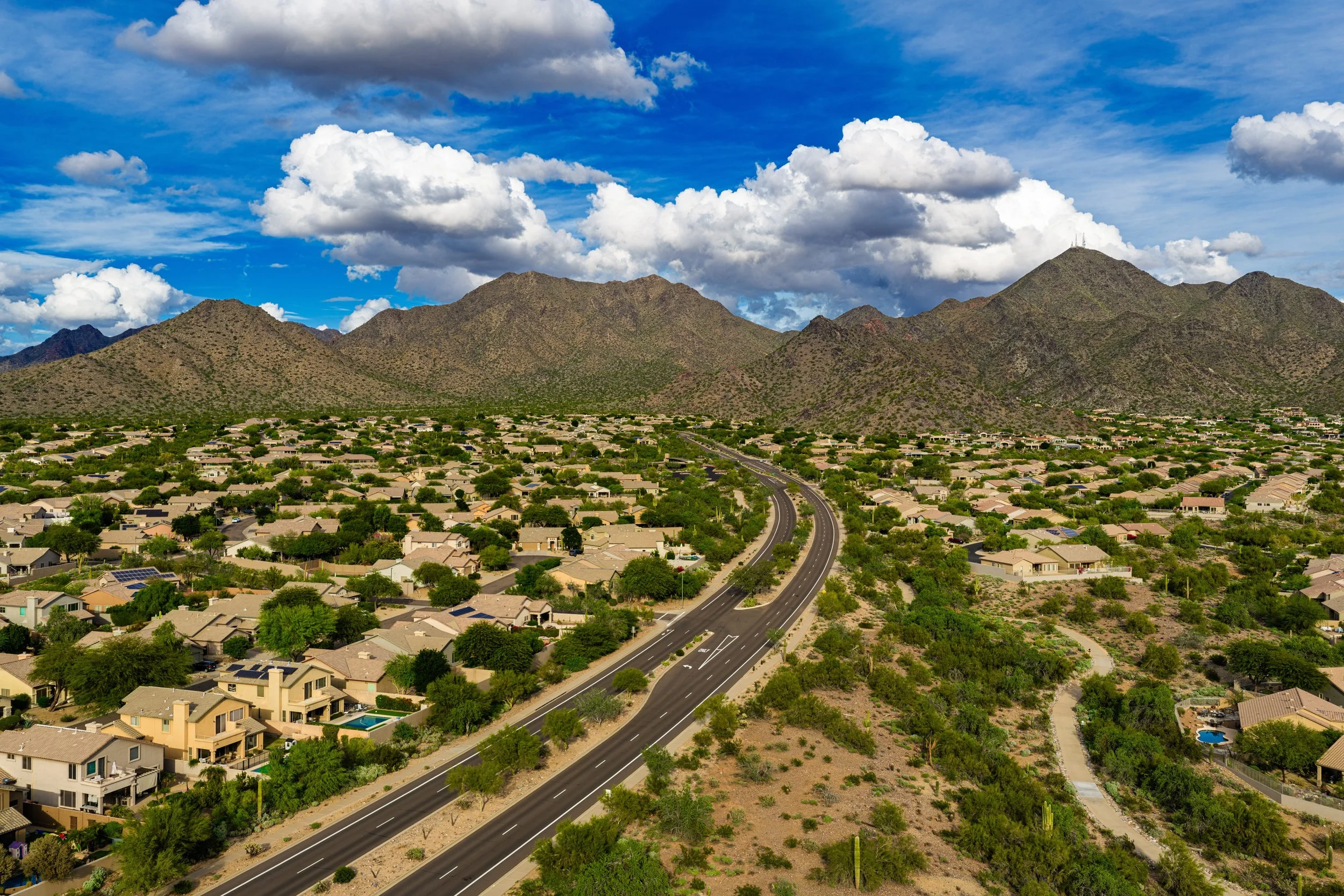 Aerial view of a suburban neighborhood with houses, trees, and a winding road, set against a mountain range under a partly cloudy blue sky.