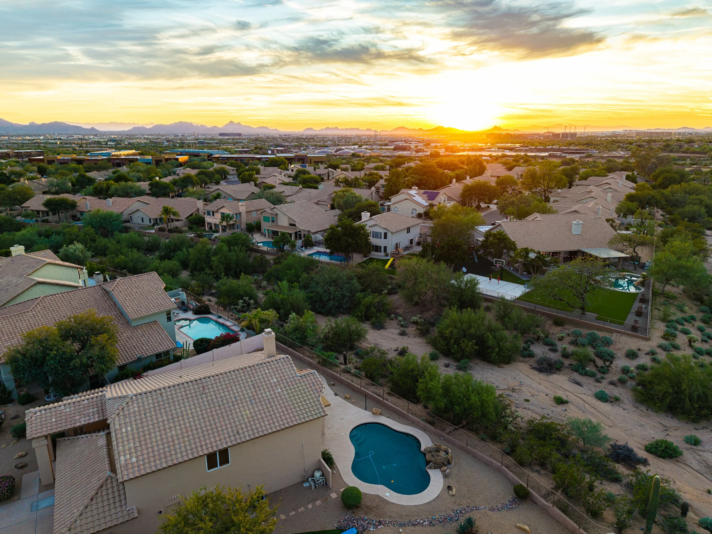 An aerial view of a residential neighborhood at sunset, showing houses with tiled roofs, swimming pools, and lush greenery including trees and bushes across a desert landscape.