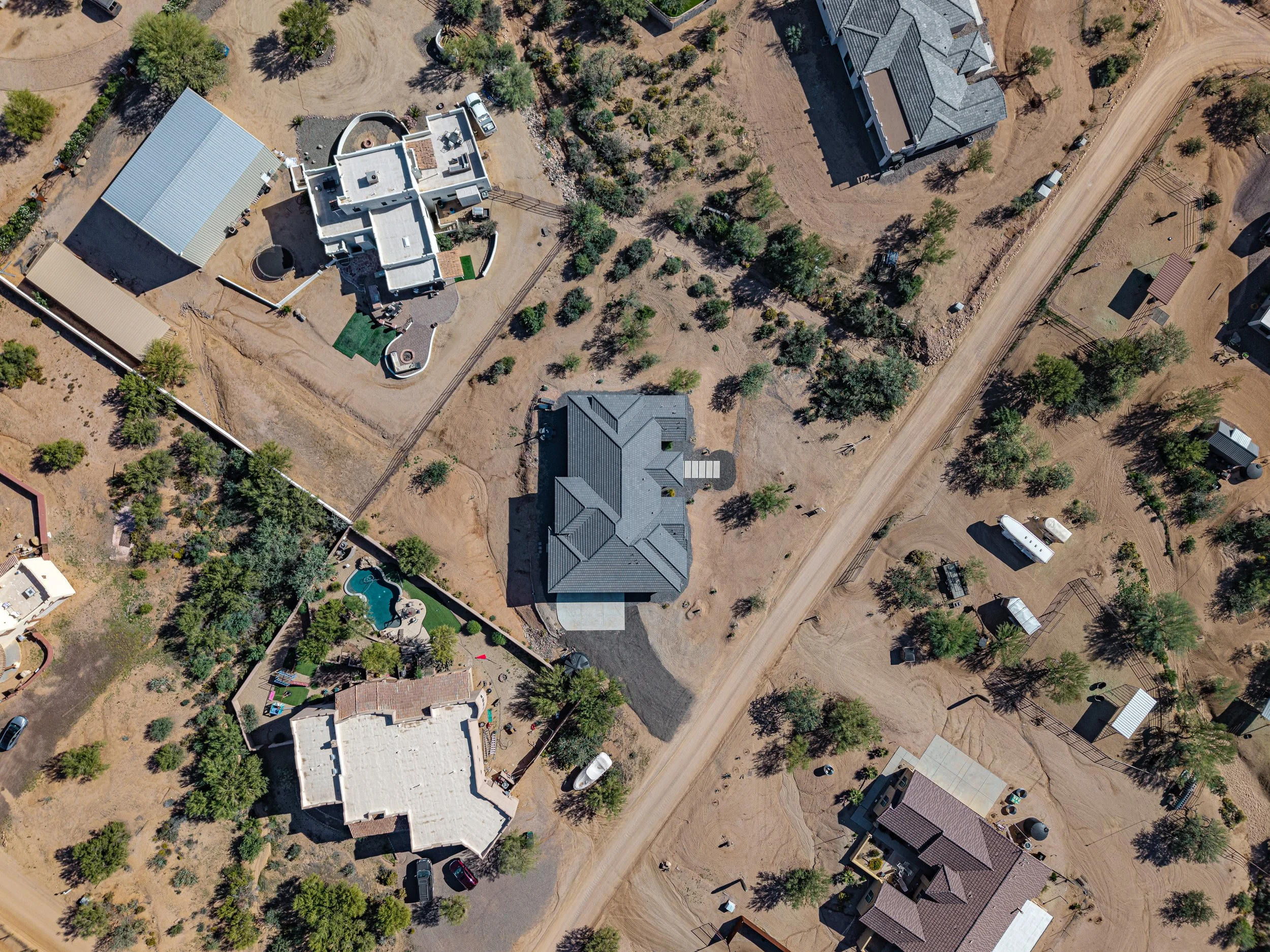 Aerial view of multiple houses in a desert-like landscape with trees, dirt roads, and outdoor features such as pools and sheds.