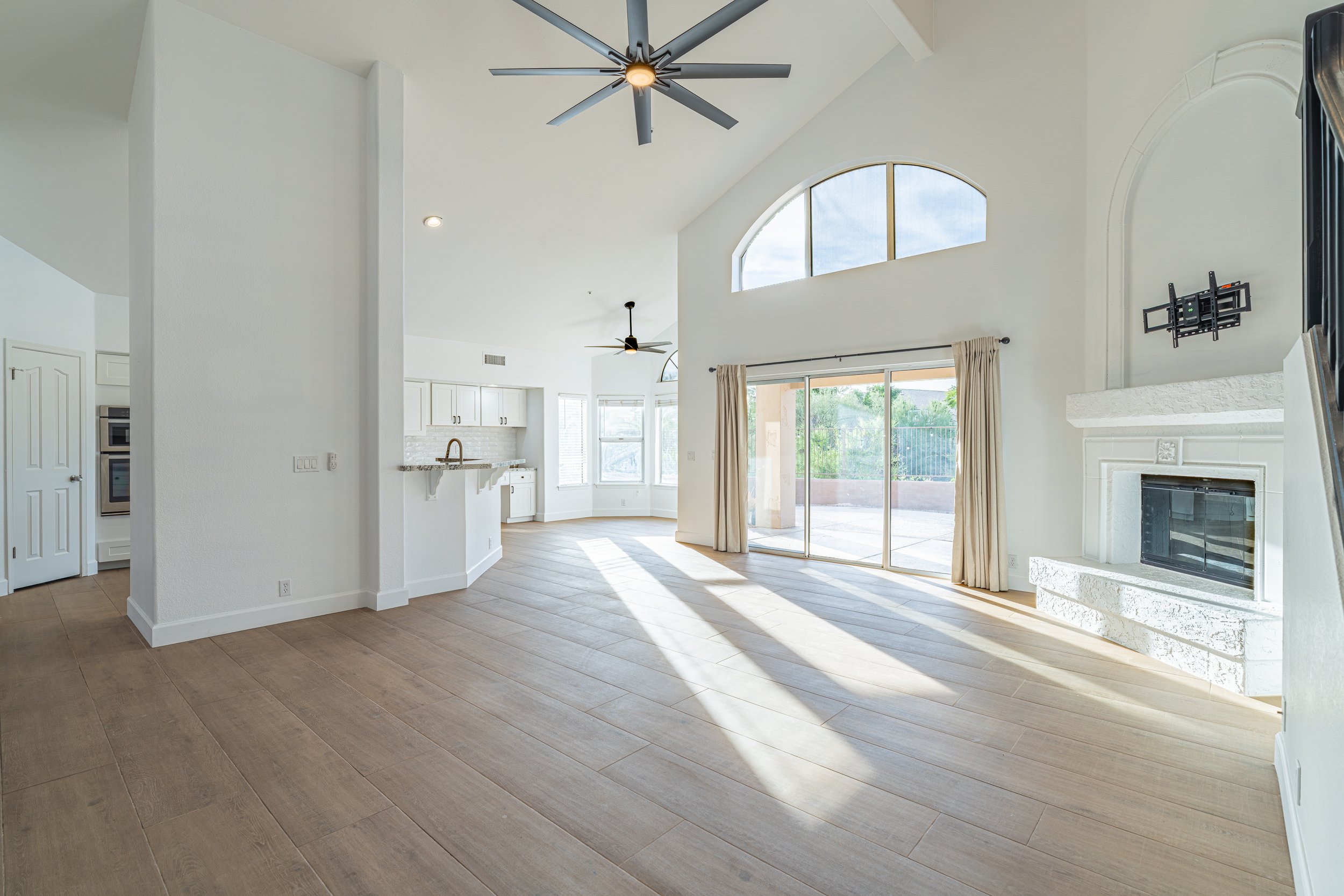 Bright, spacious living room with white walls and ceiling, large arched window, sliding glass door, and light wood flooring. Features a ceiling fan, fireplace, and views of the outdoor patio.