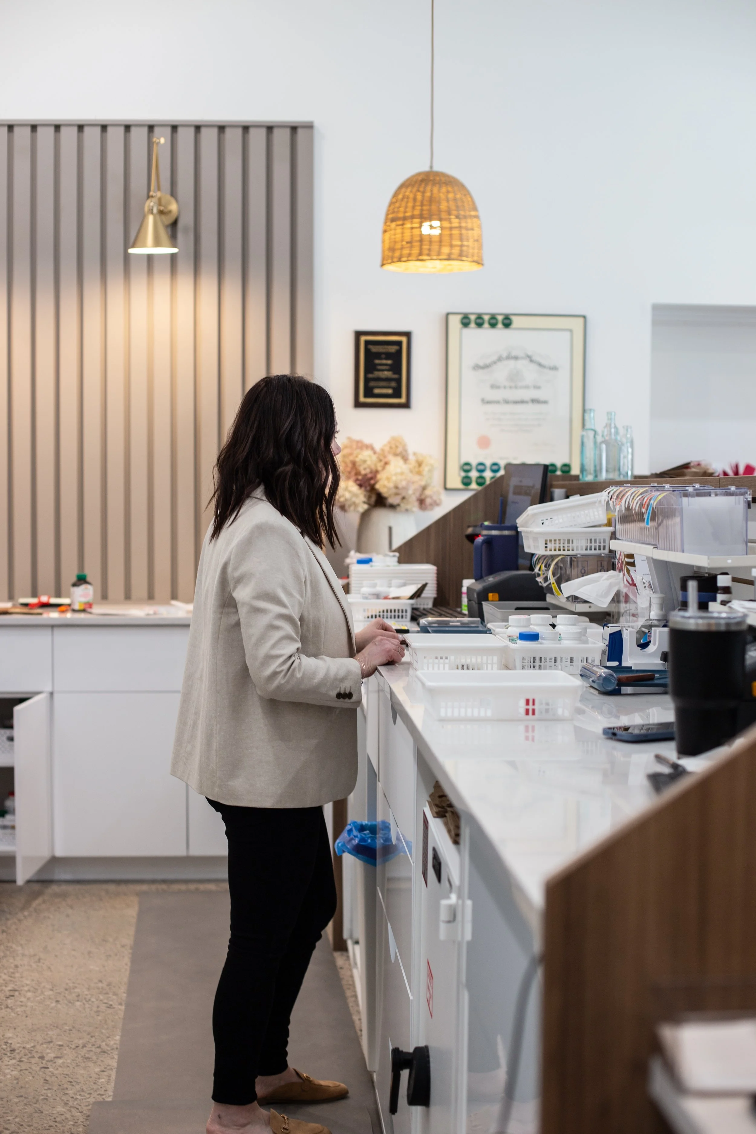Woman with dark hair wearing a beige blazer and black pants standing at a countertop with medical supplies and equipment in a pharmacy.