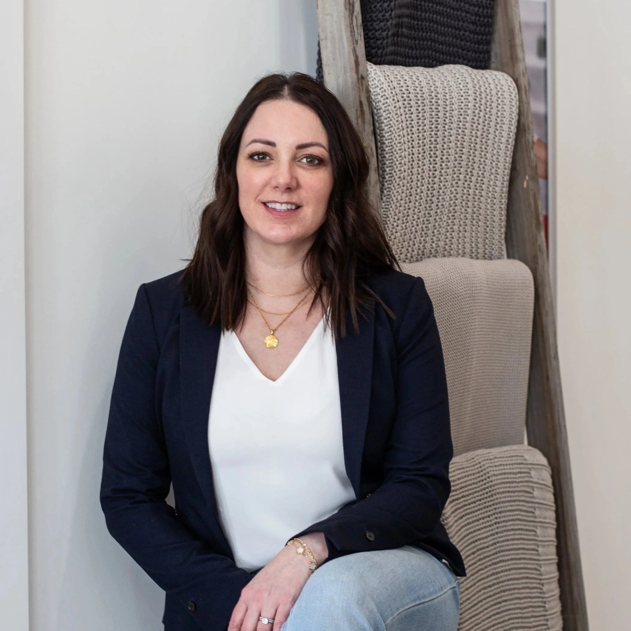 A woman with shoulder-length dark hair, wearing a black blazer, white top, and light-colored jeans, sitting on a chair in front of a wooden display with different textured fabric samples. She is smiling and accessorized with gold jewelry.