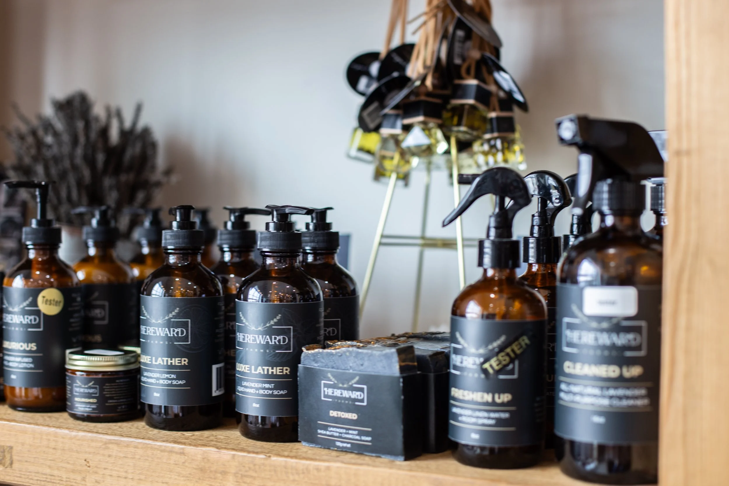 A display of brown bottles with black labels and pump dispensers on a wooden shelf, with some soaps labeled 'Luxe Lather' and 'Refresh Up' and a bar of soap in a box, along with a decorative halo of small bottles in the background.
