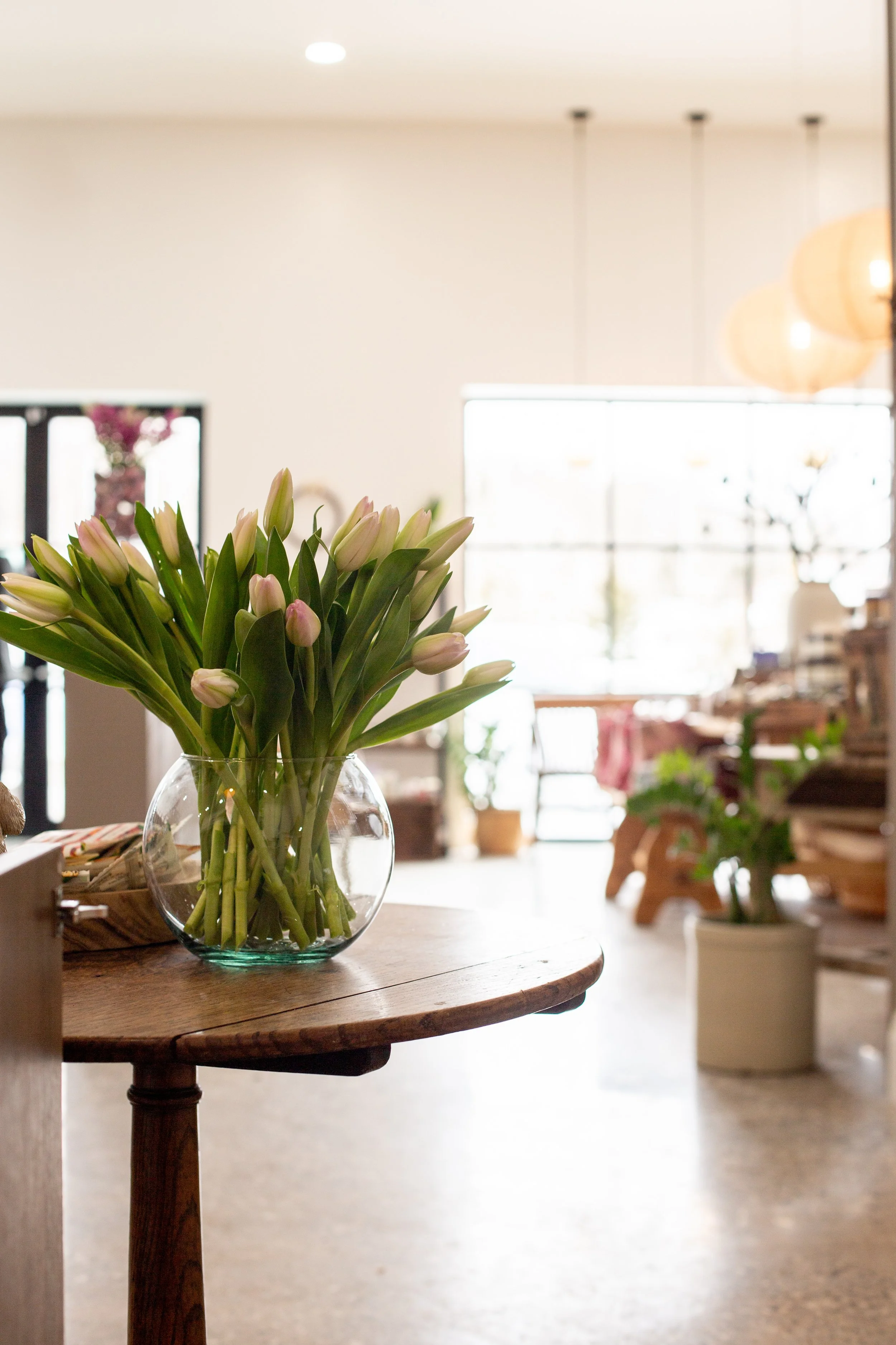 A glass vase with white and pale pink tulips on a round wooden table in a bright, modern room with large windows, indoor plants, and hanging lights.