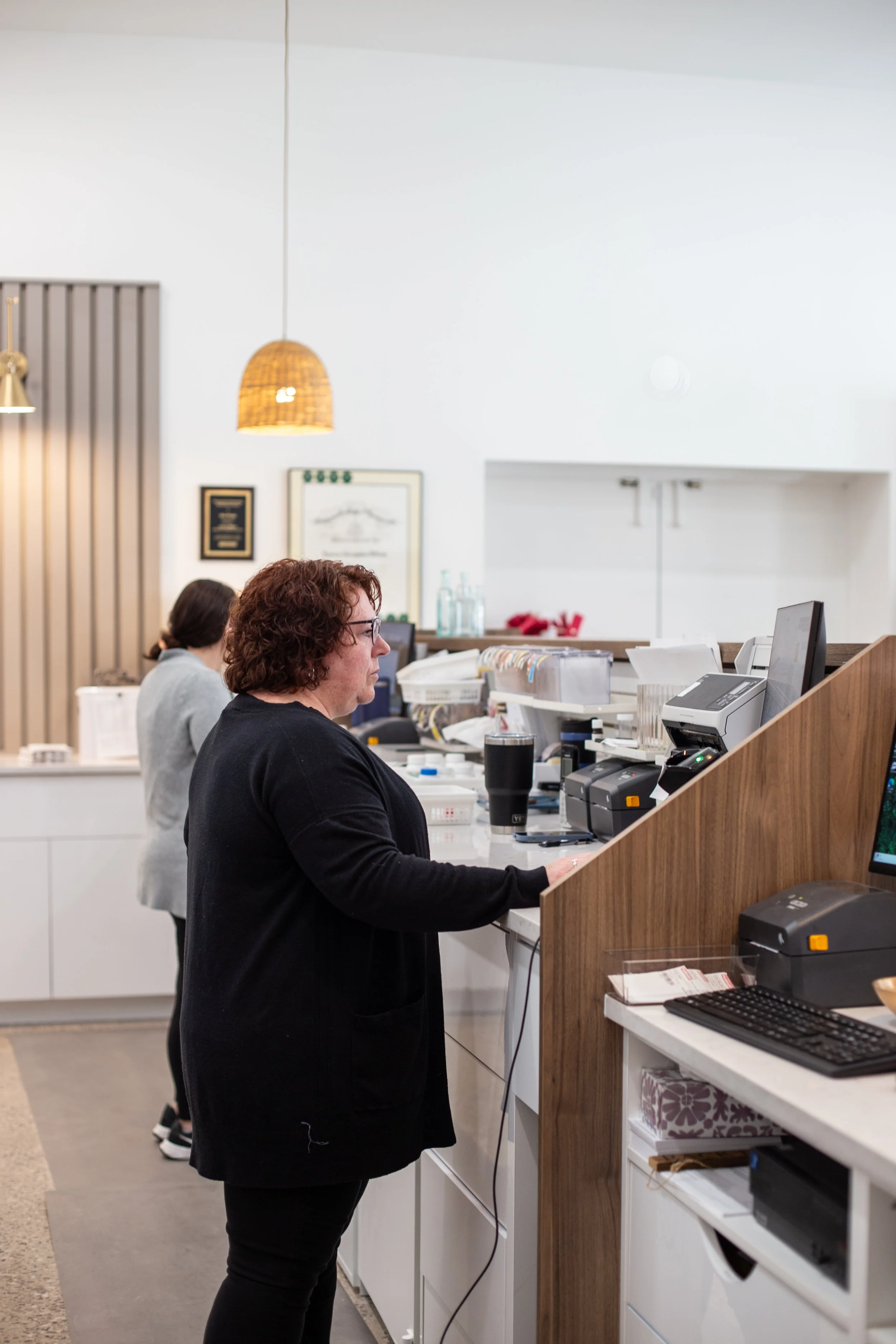 A woman with curly hair wearing glasses and a black jacket standing at a reception desk in a pharmacy, with another woman in the background working at a counter.