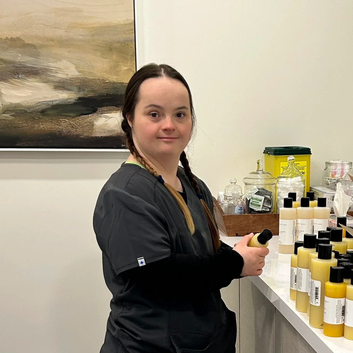 Young woman with braided hair standing at a pharmacy counter, holding a yellow bottle with a black cap, surrounded by bottles of similar size on the counter.