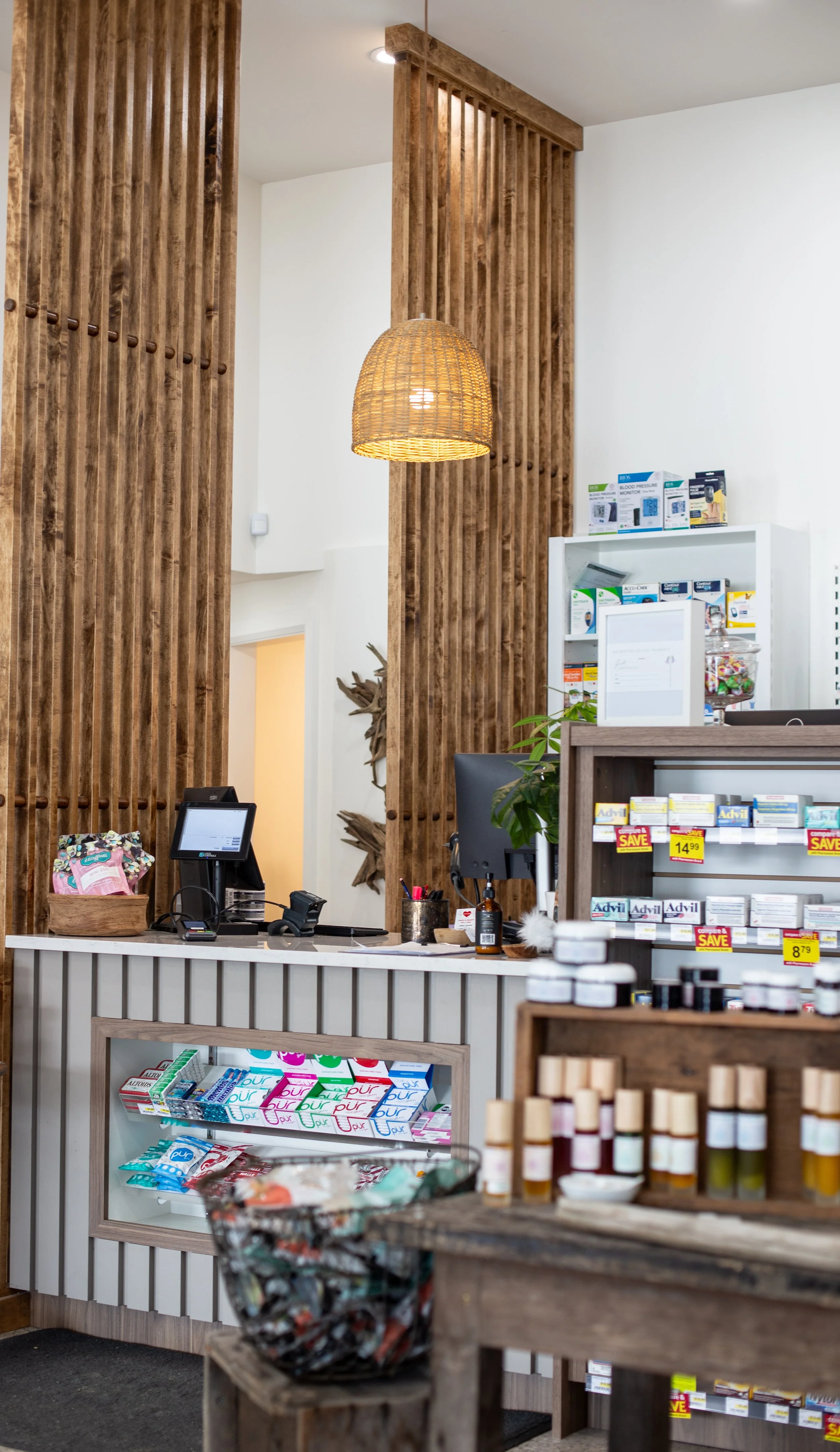 Interior of a pharmacy showing a wooden counter, a computer, shelves with medication, and a decorative wooden slat partition with a hanging woven light fixture.