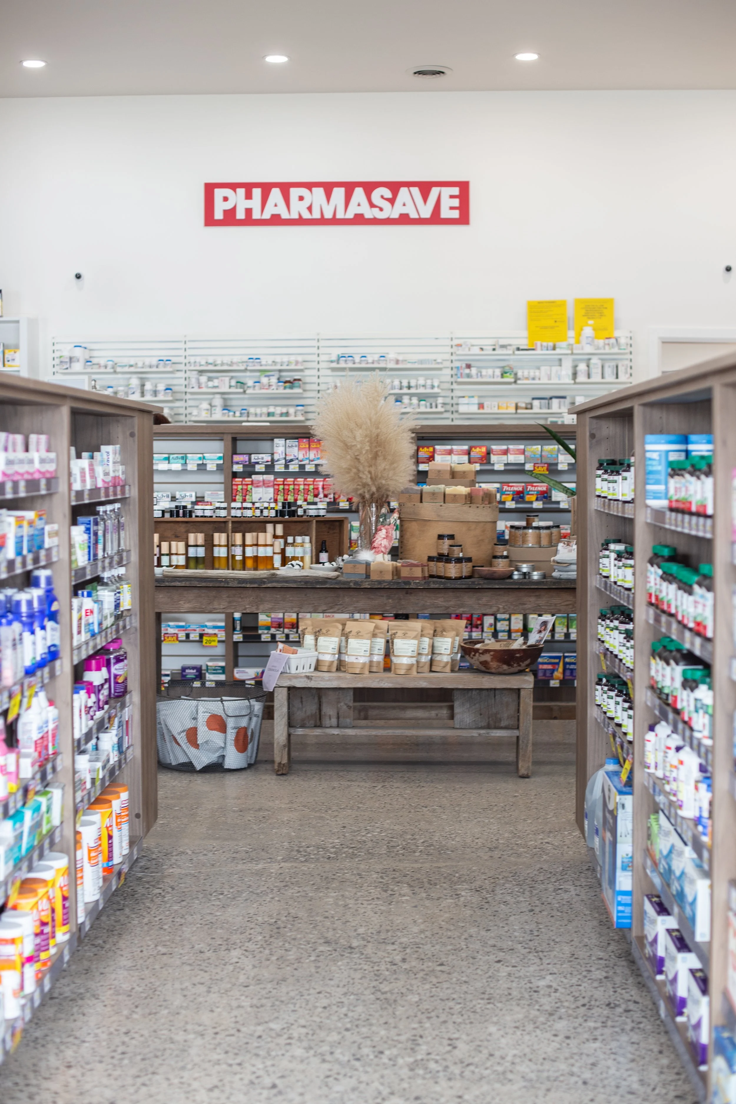Interior of a pharmacy, with shelves of medicine and health products on both sides, a display table in the center with natural decor, and a large red sign reading 'PHARMASAVE' on the back wall.
