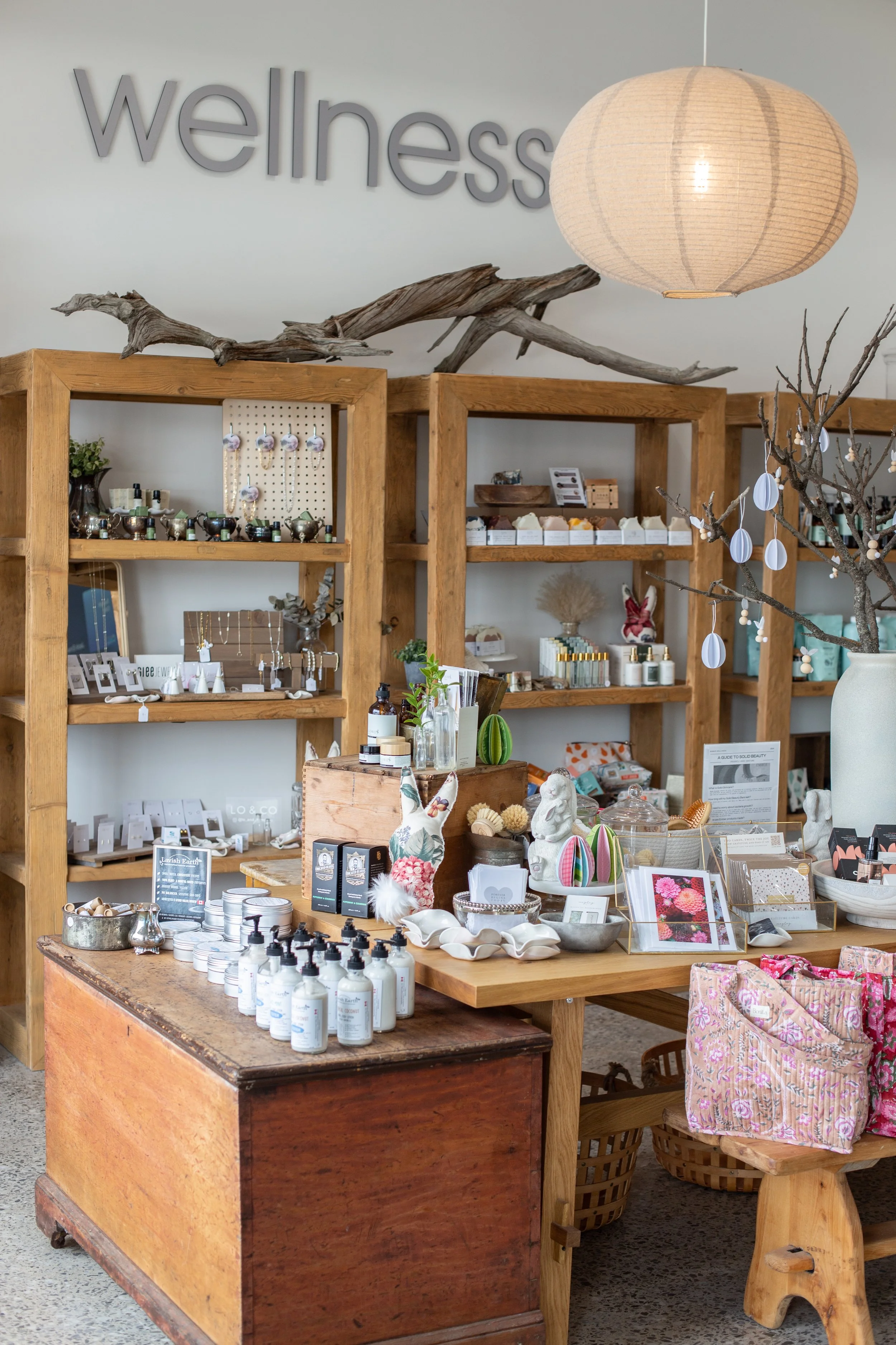 Display of wellness and body care products on wooden shelves and tables in a retail store with a sign reading 'wellness' on the wall.