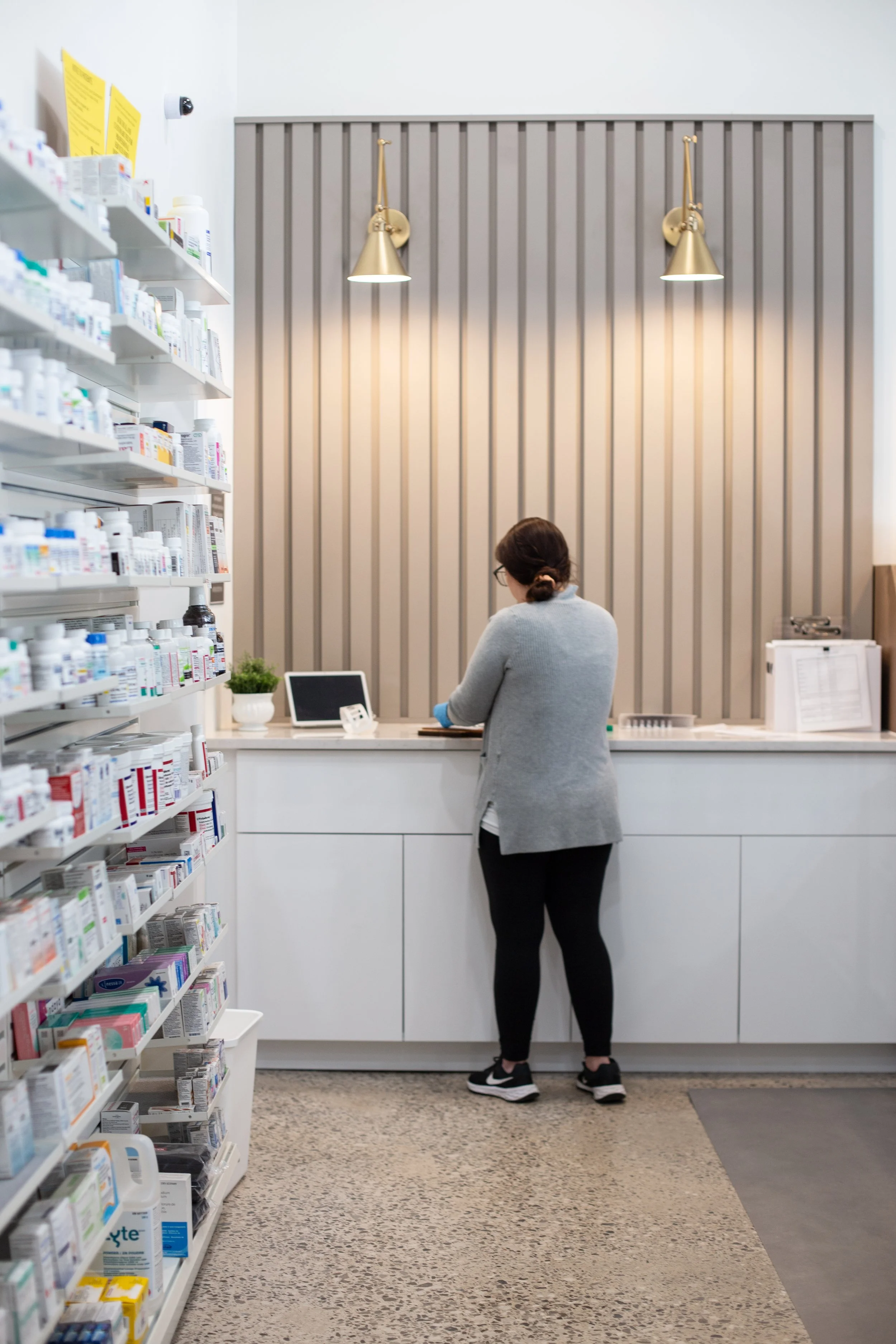 A woman in a gray sweater and black leggings standing at a pharmacy counter with a tablet device, shelves full of medication on the side, in a modern pharmacy setting.