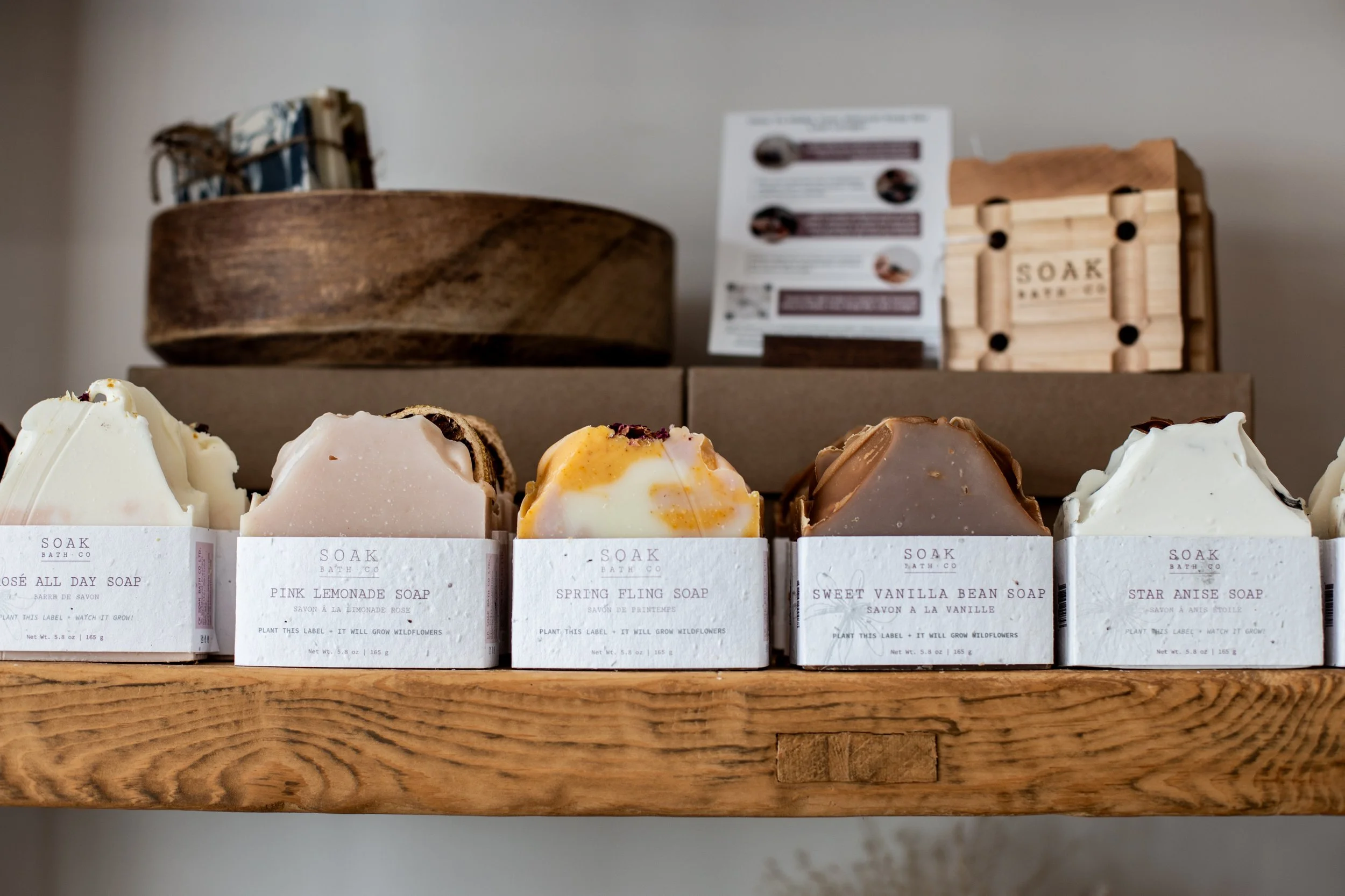Display of five soap bars with white labels on a wooden shelf, with a round wooden bowl and a wooden soap holder in the background.
