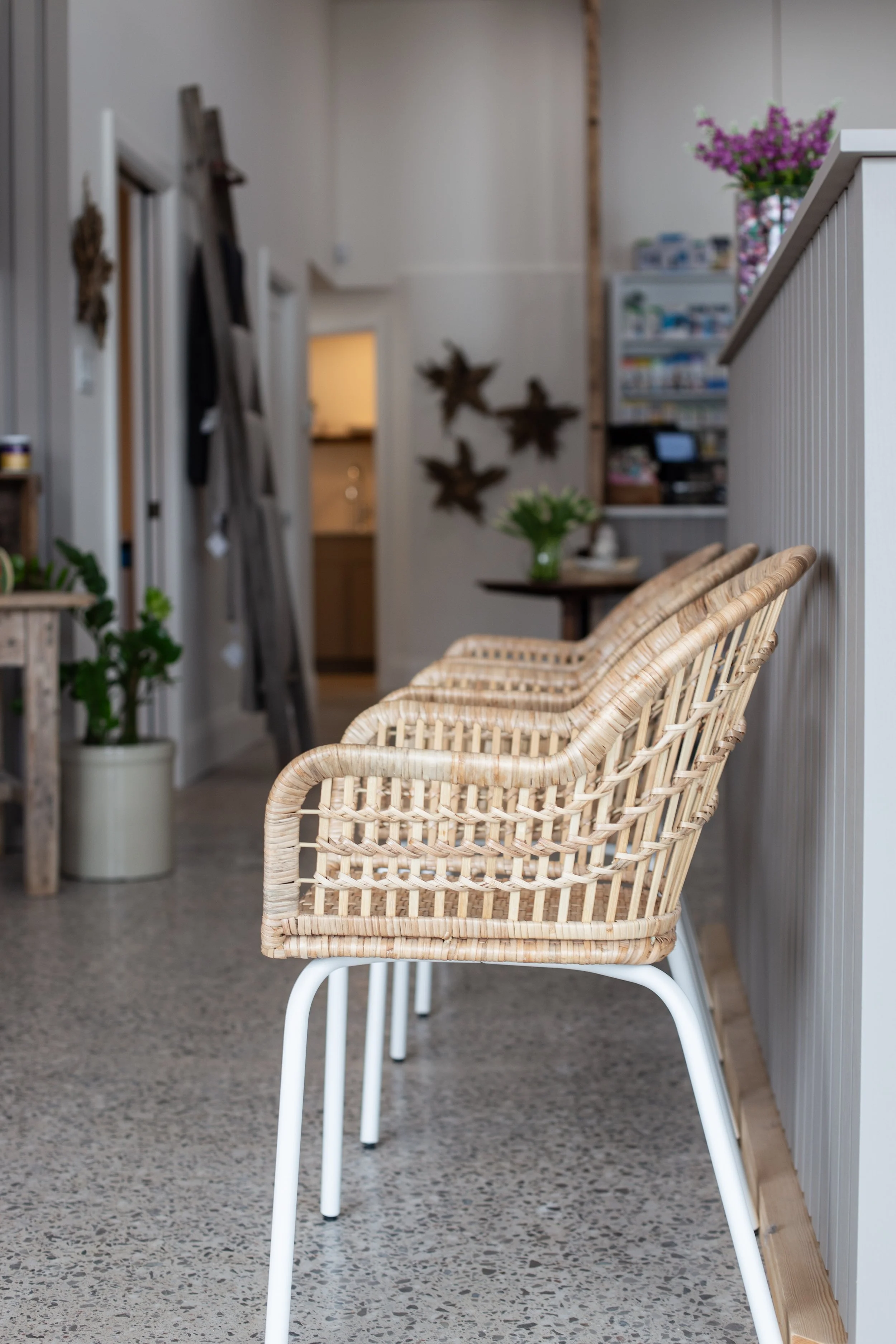 Three wicker chairs with white metal legs are lined up next to a white-paneled half wall in a cozy interior space. In the background, there is a table with a green vase of white flowers and a shelf with various items. A potted plant is on the floor to the left, and there are decorative wall hangings and a ladder leaning against the wall.