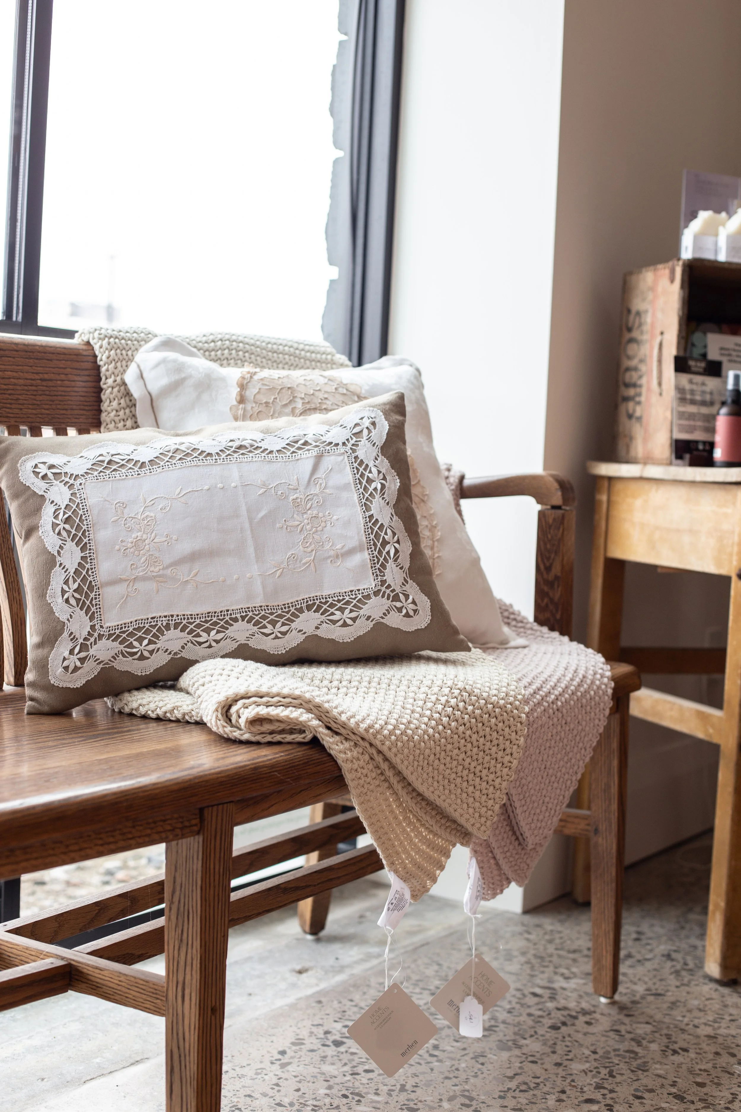 A wooden bench with neutral-colored embroidered pillows and a knitted blanket, placed in front of a window with sunlight streaming in.