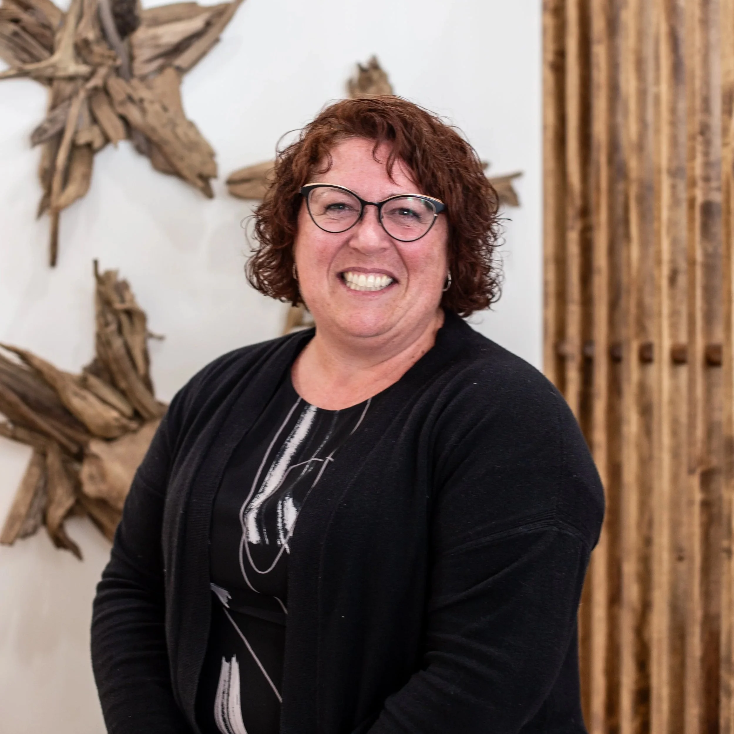 A woman with curly red hair and glasses smiling indoors, with wooden art on the wall behind her.