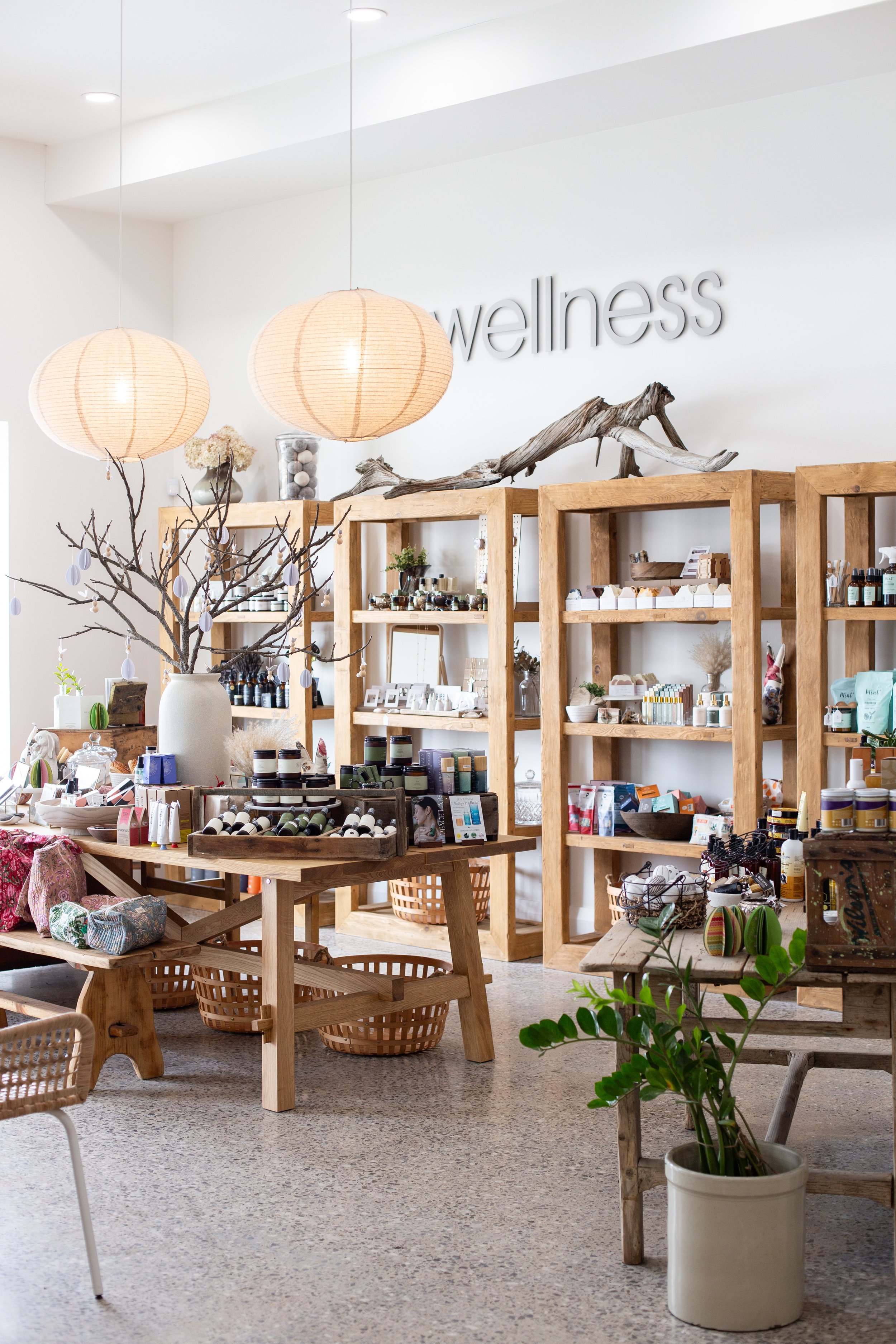 Interior of a wellness shop with wooden shelves displaying natural products, a wooden table with botanical items, paper lantern lighting, and decorative elements like branches and plants, with the word 'wellness' on the wall.