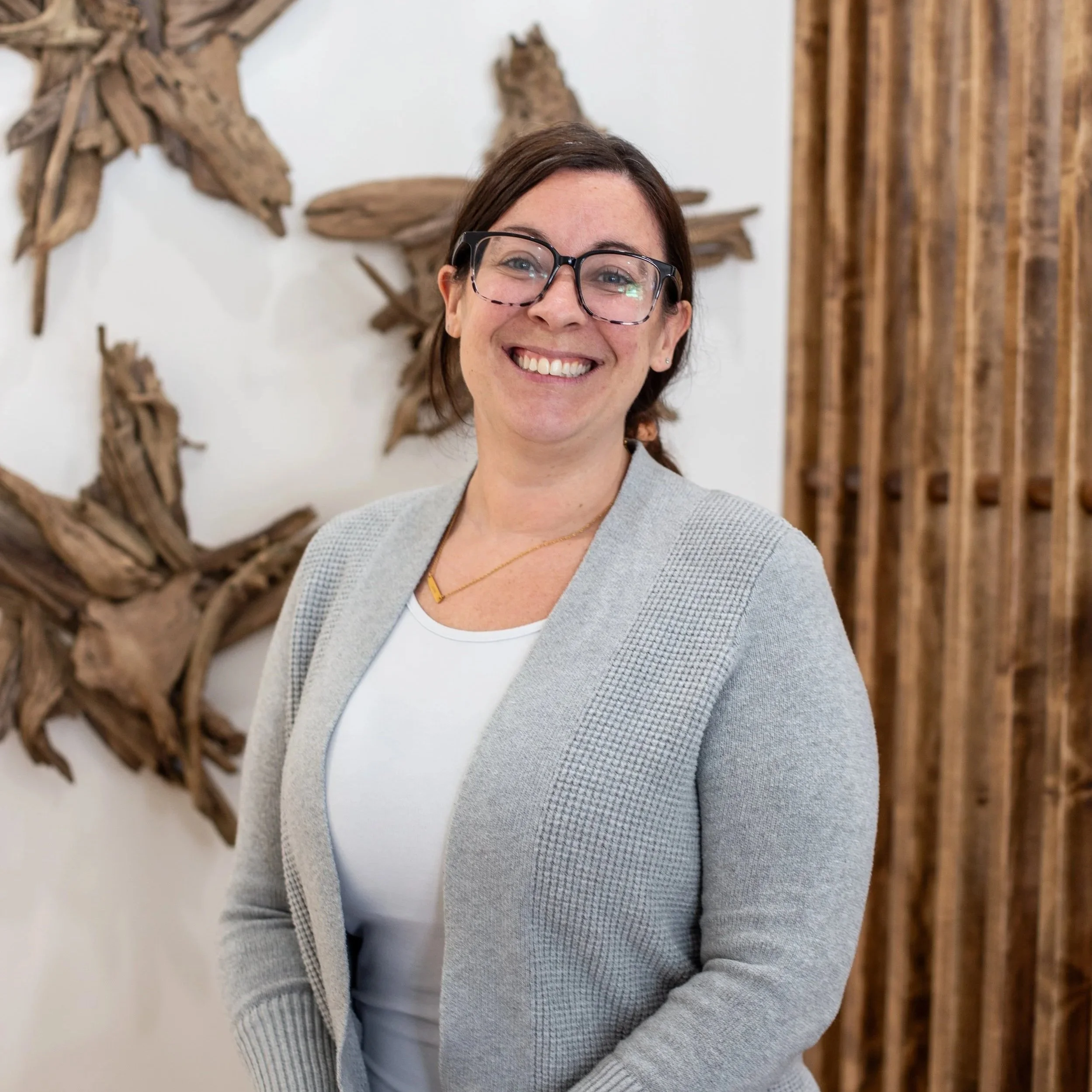 A woman with glasses smiling in a professional setting with wooden art on the wall.