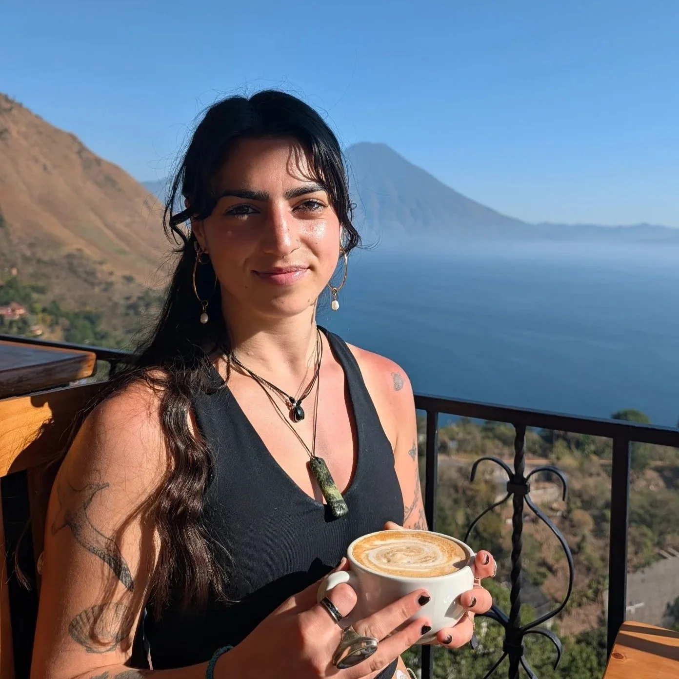 A woman with dark hair is holding a cup of coffee, standing on a balcony overlooking a body of water and mountains.