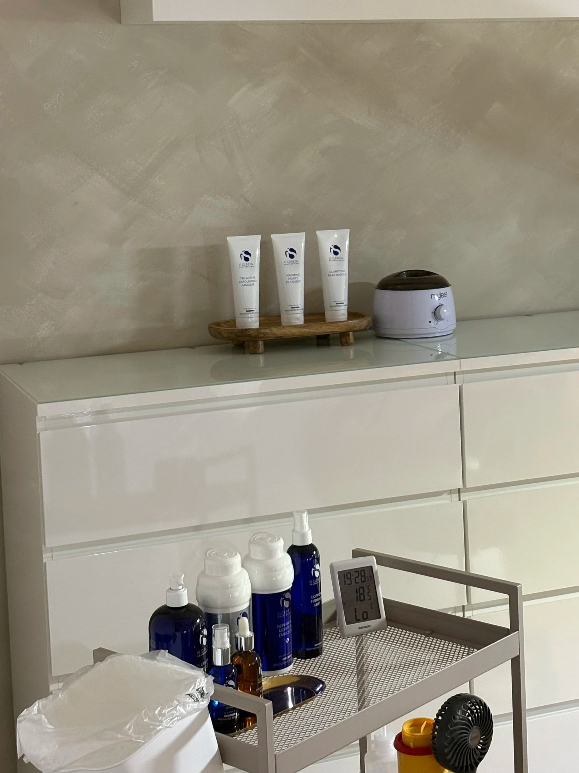 Eine weiße Kommode mit verschiedenen Pflegeprodukten auf einem kleinen Wagen davor, einem Digitalthermometer und einem Ventilator.