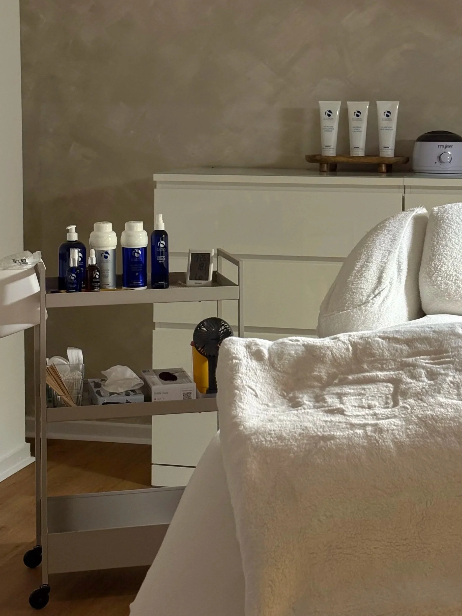 Ein Raum mit weißen Möbeln, ein Regal mit Pflegeprodukten, einem weißen Handtuch, einem kleinen schwarzen Ventilator und einem digitalen Thermometer, die entspannte Atmosphäre suggerieren.