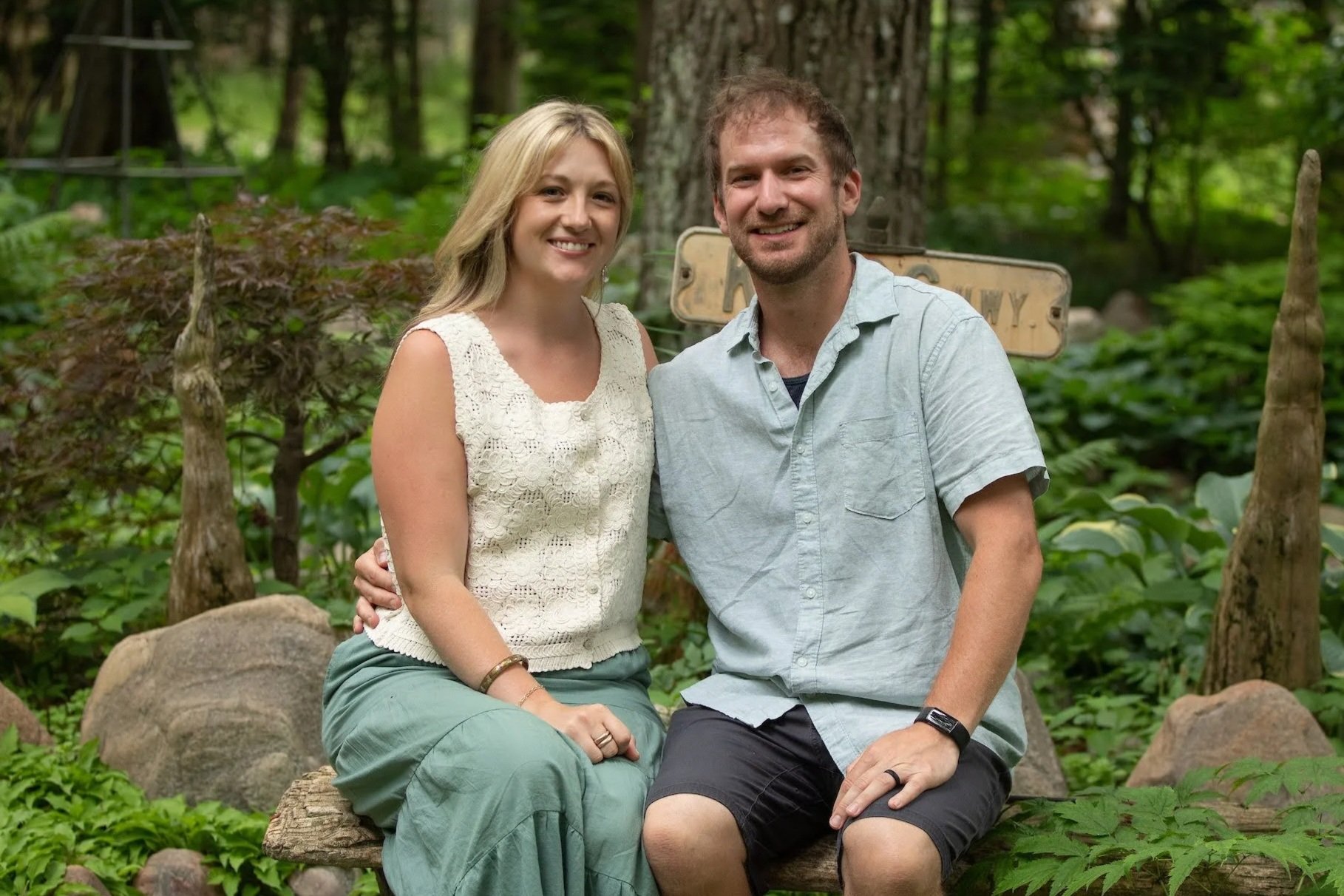 A smiling couple sitting on a wooden bench in a lush green forest, with a sign behind them and surrounded by trees and plants.