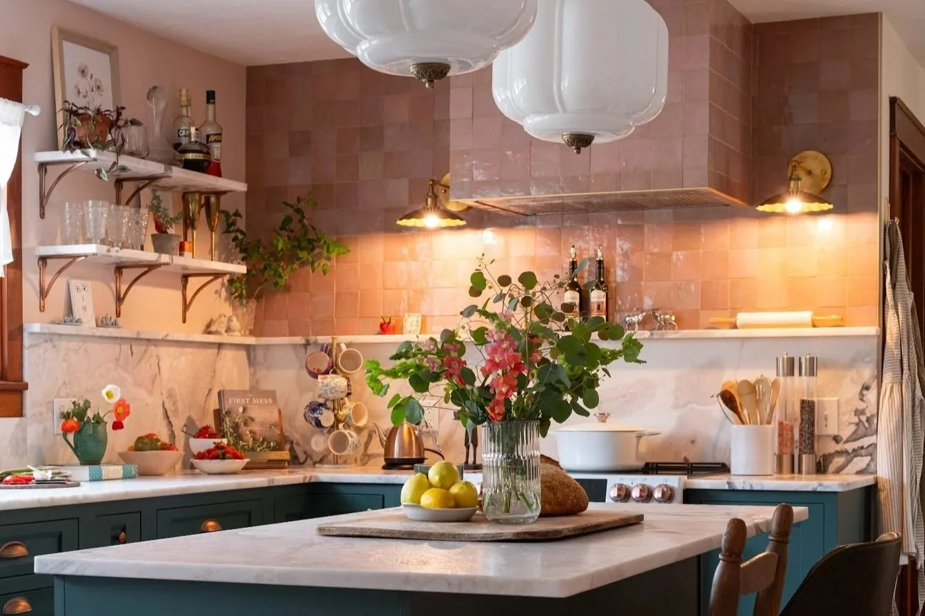 Cozy kitchen with pink tiled walls, white marble countertops, and teal cabinets. Decorated with a flower vase, bowls of fruit, books, and hanging mugs. Open shelves with glasses and bottles, and soft lighting.