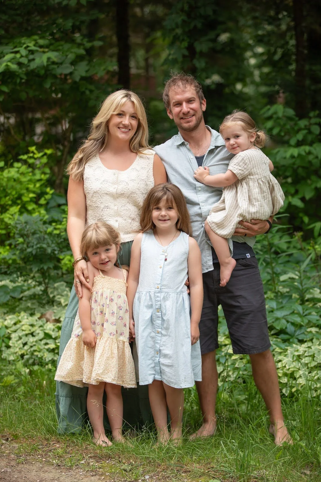 A family of six standing outdoors in a wooded area, smiling at the camera. The group includes two adults and four young children, with greenery and trees in the background.