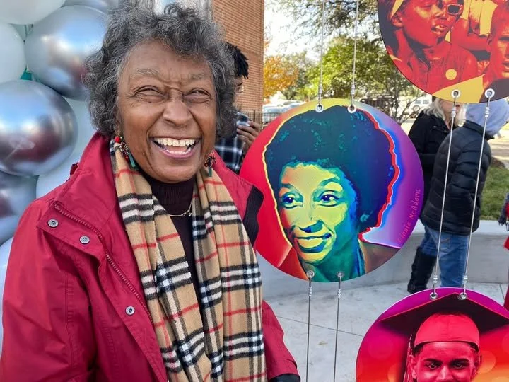 Linnie McAdams stands outdoors, smiling warmly next to a colorful circular portrait of herself featured in the Denton Women’s Interracial Fellowship monument at Industrial Street Park.