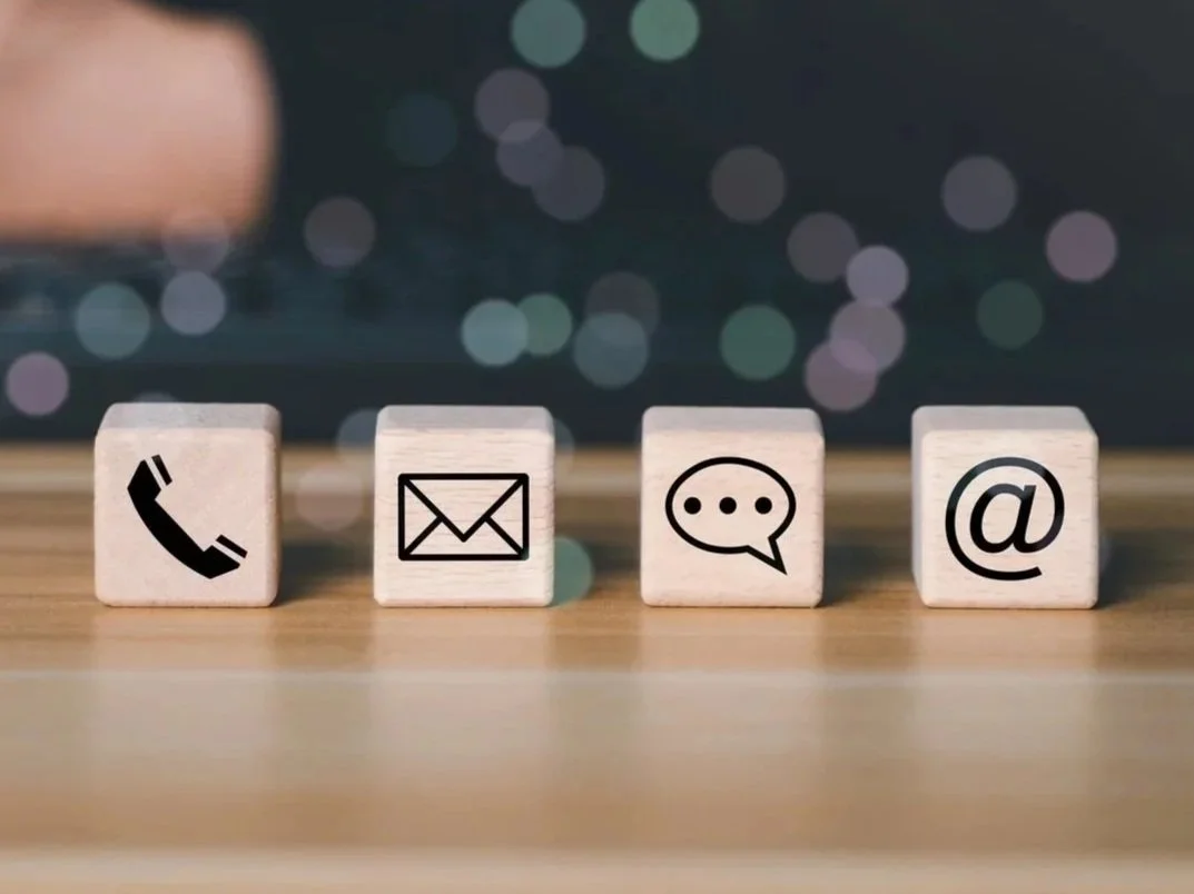 Four wooden blocks with icons of a phone, an envelope, a speech bubble, and an at symbol, arranged on a wooden surface.
