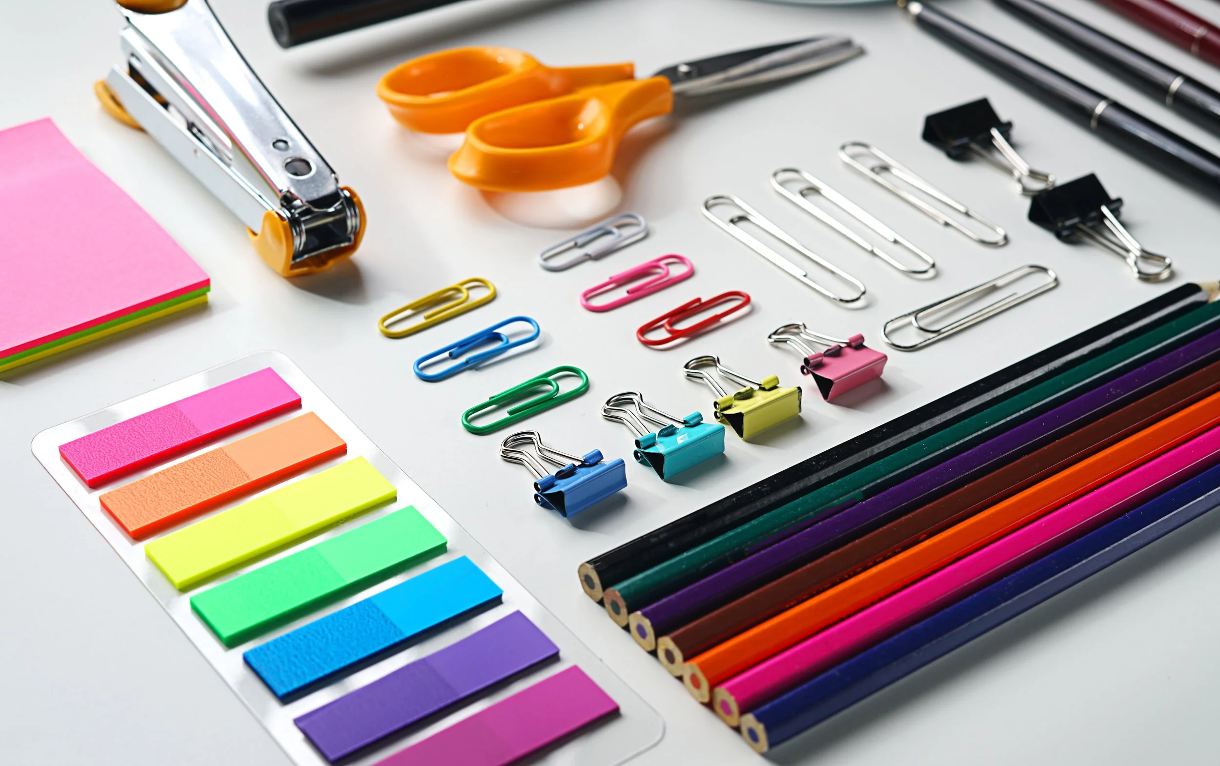Office supplies including scissors, binder clips, paper clips, colored pencils, sticky notes, and pens on a white desk.