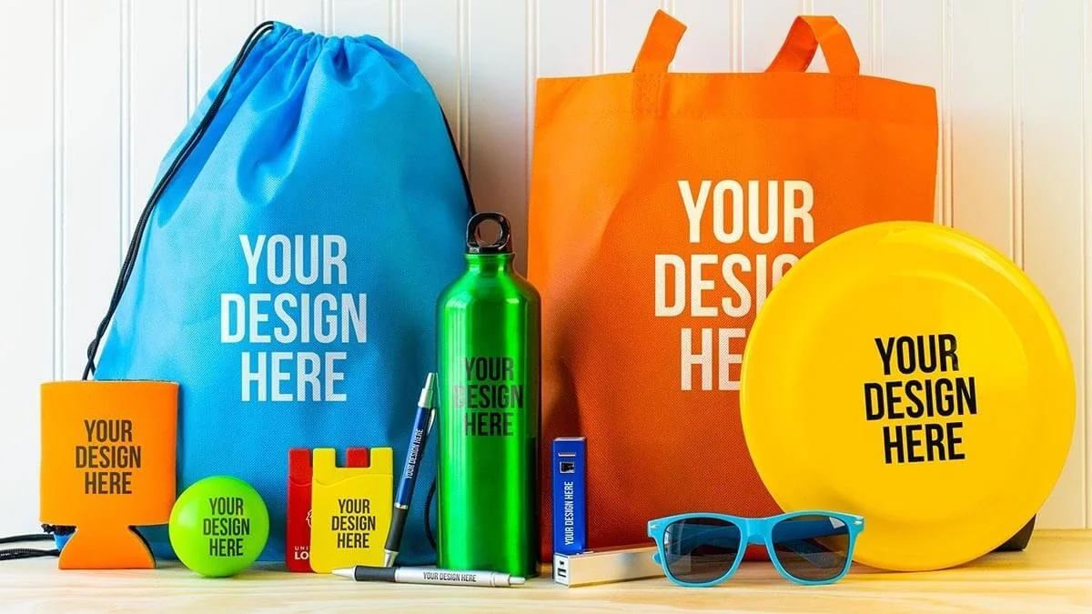 Collection of customizable items including drawstring bags, tote bag, frisbee, water bottle, foam toy, notebook, pen, and sunglasses, all with the placeholder text "Your Design Here" on brightly colored backgrounds.