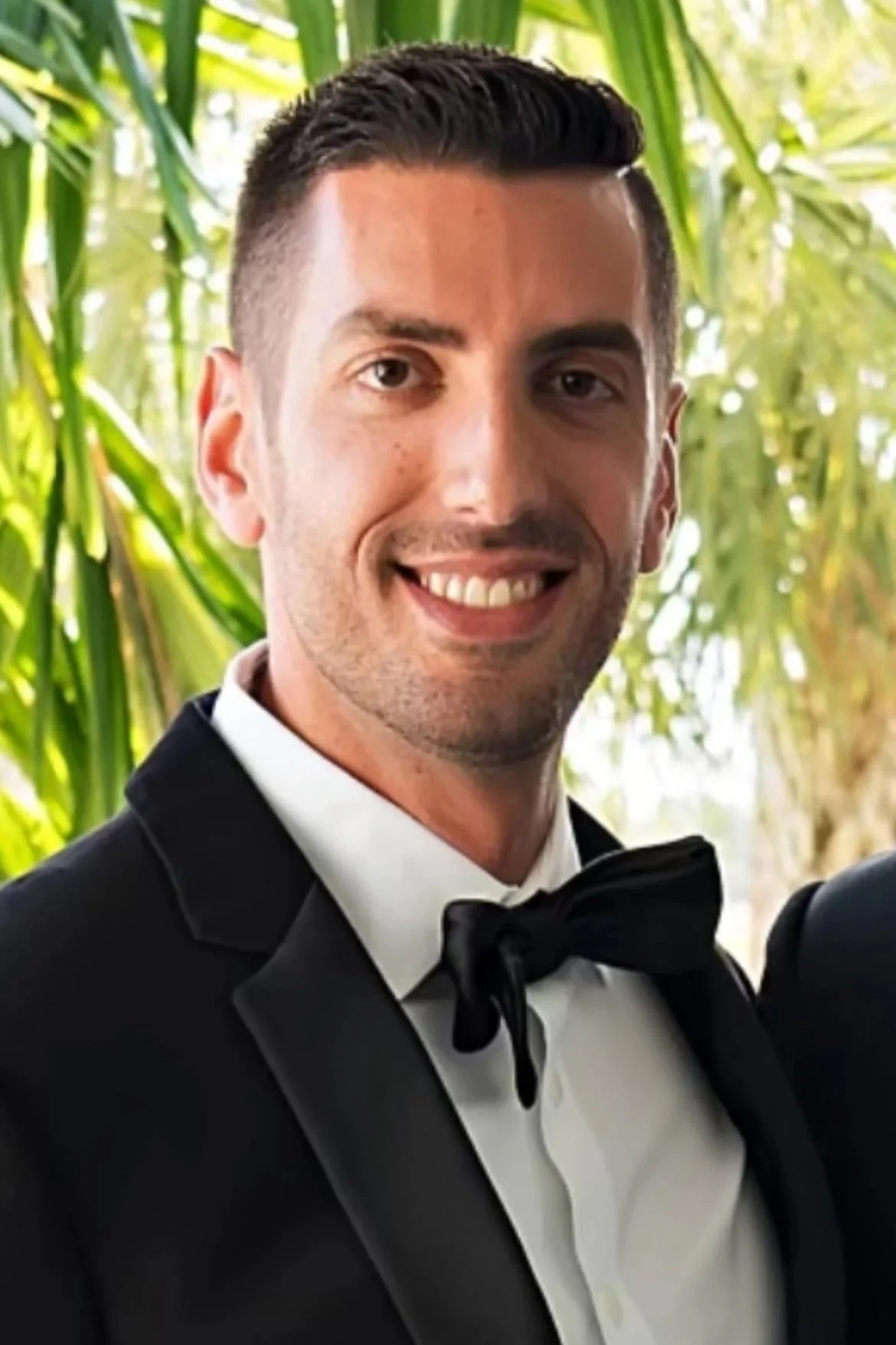 A man in a tuxedo with a bow tie smiling, with green leafy plants in the background.