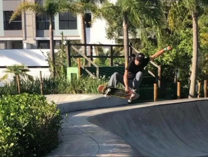 Skatista fazendo manobra em skatepark ao ar livre, cercado por árvores e plantas.