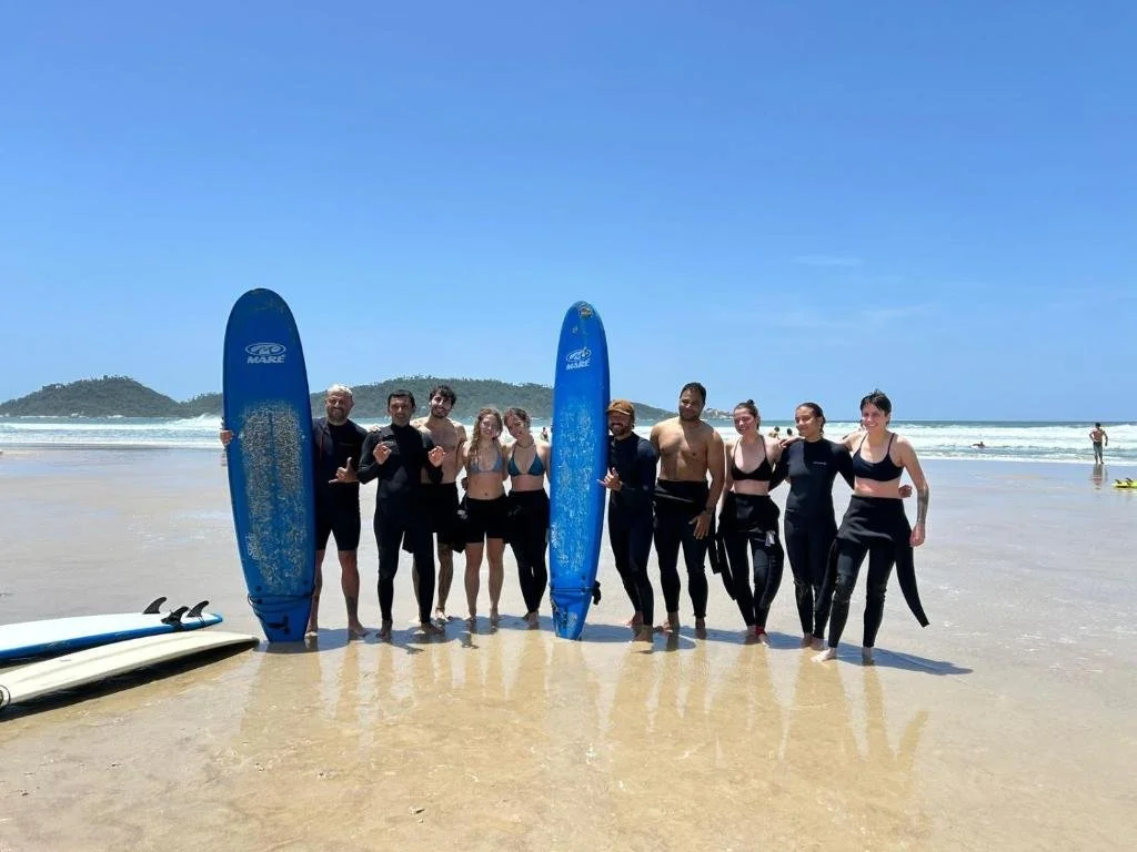 Grupo de pessoas na praia com pranchas de surf, ao fundo mar, céu azul e ilhas ao longe.