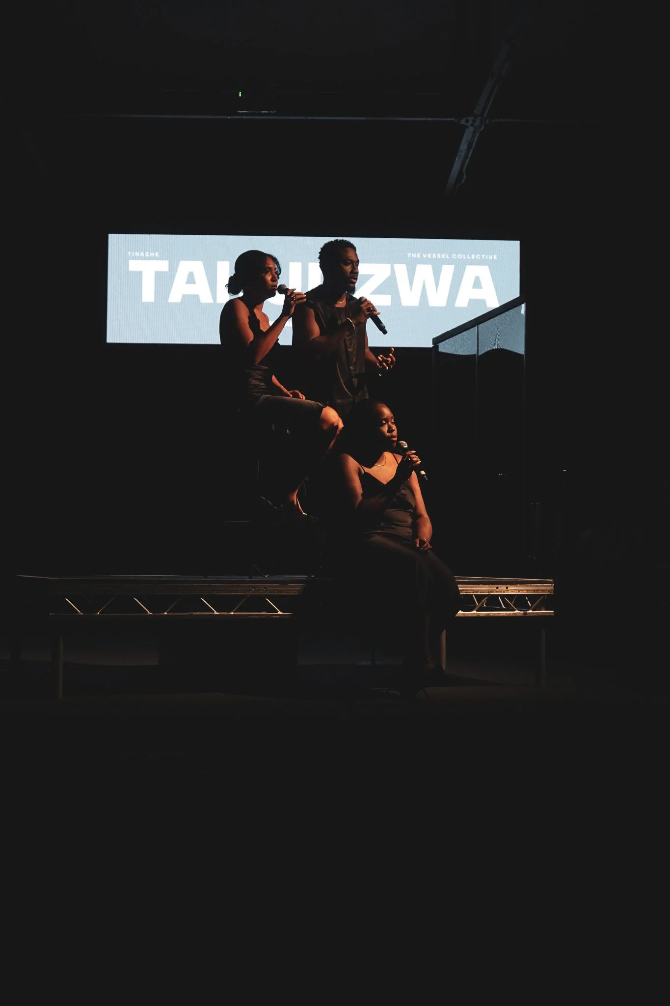 Three people are on stage, holding microphones, with a large screen behind them displaying the text 'TASIM ZWA' and 'The Vessel Collective'. The photo is dark, with the individuals illuminated by the stage lighting.