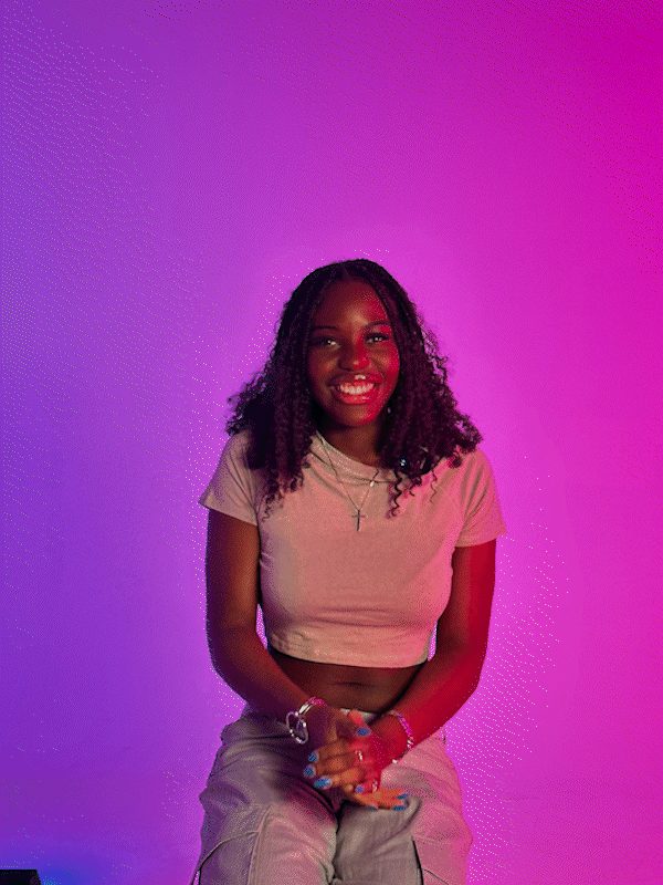 A young woman with curly hair, wearing a beige crop top, sitting against a pink and purple gradient background, smiling at the camera.
