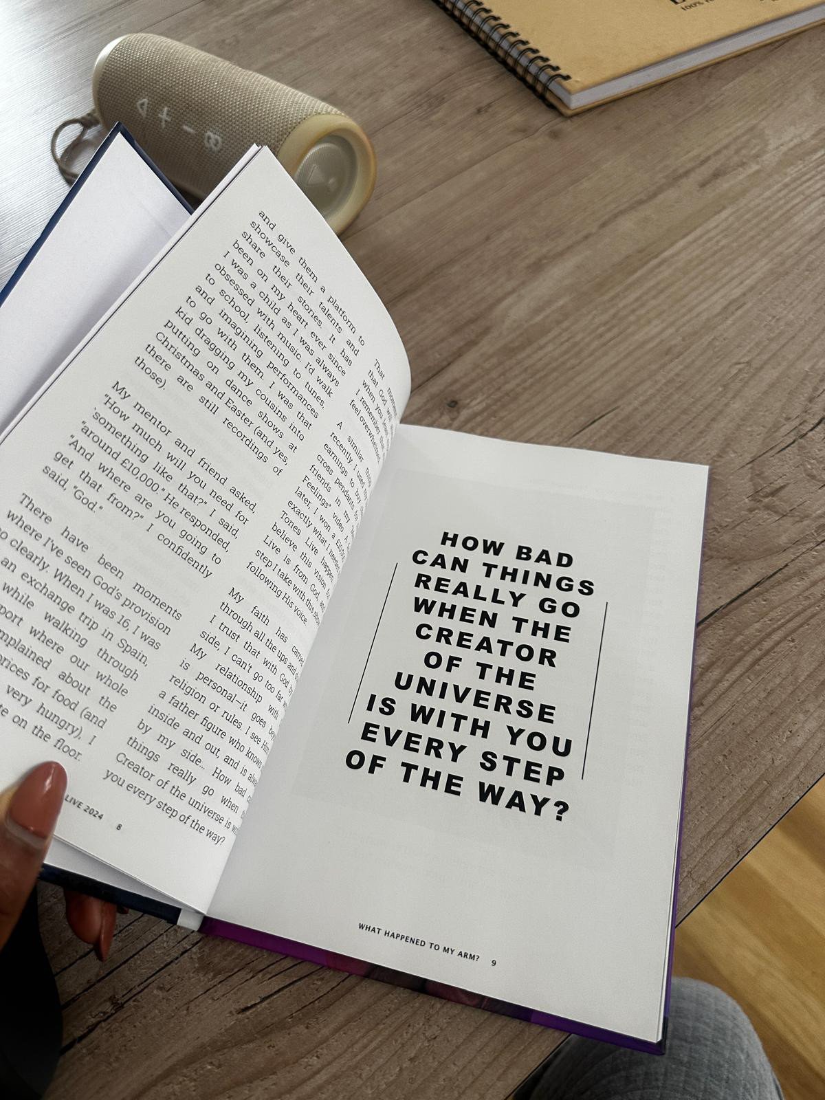 Open book on a wooden table with a bold quote that reads: "How bad things can truly go when the creator of the universe is with you every step of the way?"; a portable speaker and another closed book can be seen nearby.